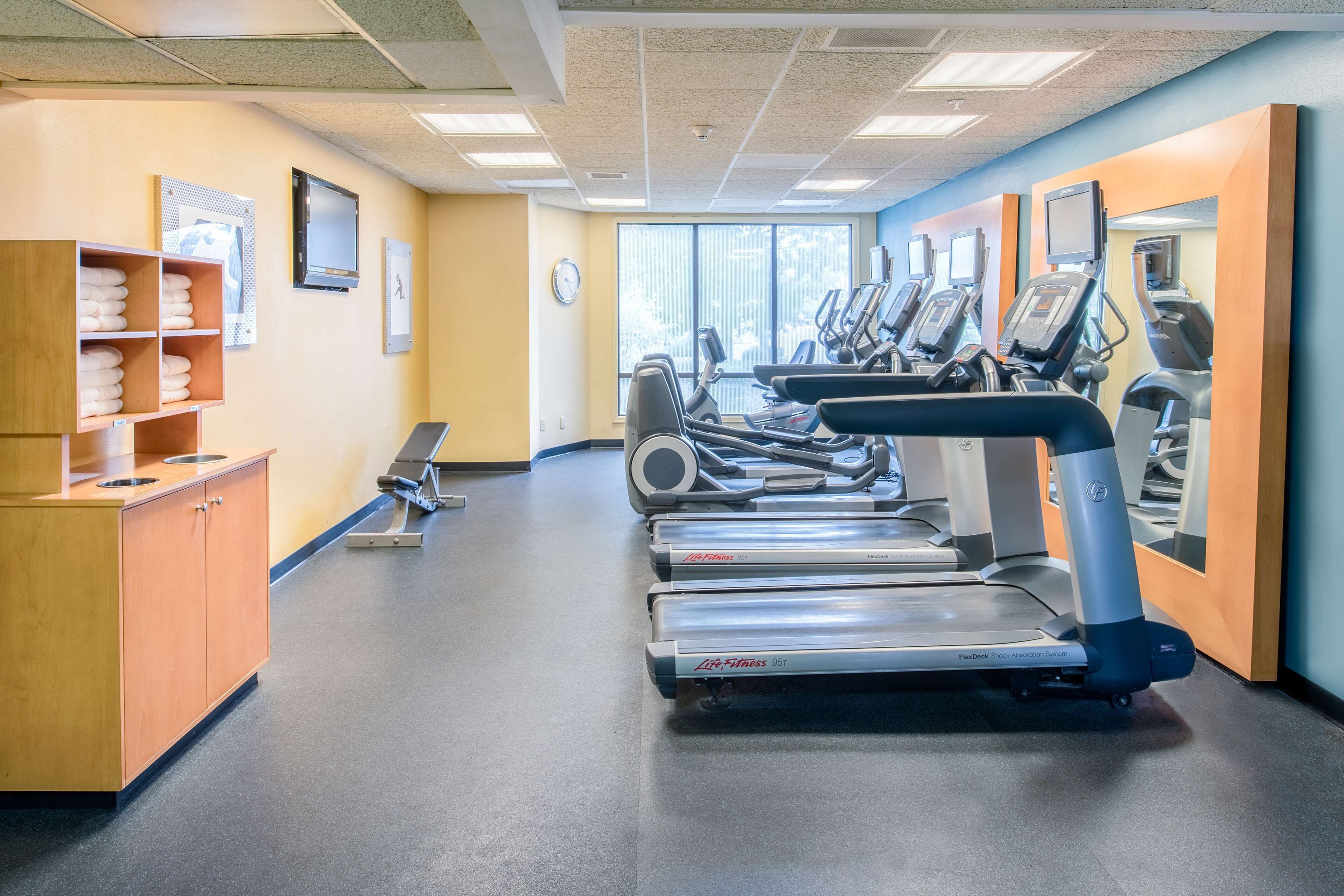 A gym with treadmills, a wooden cabinet with towels, and a large window.
