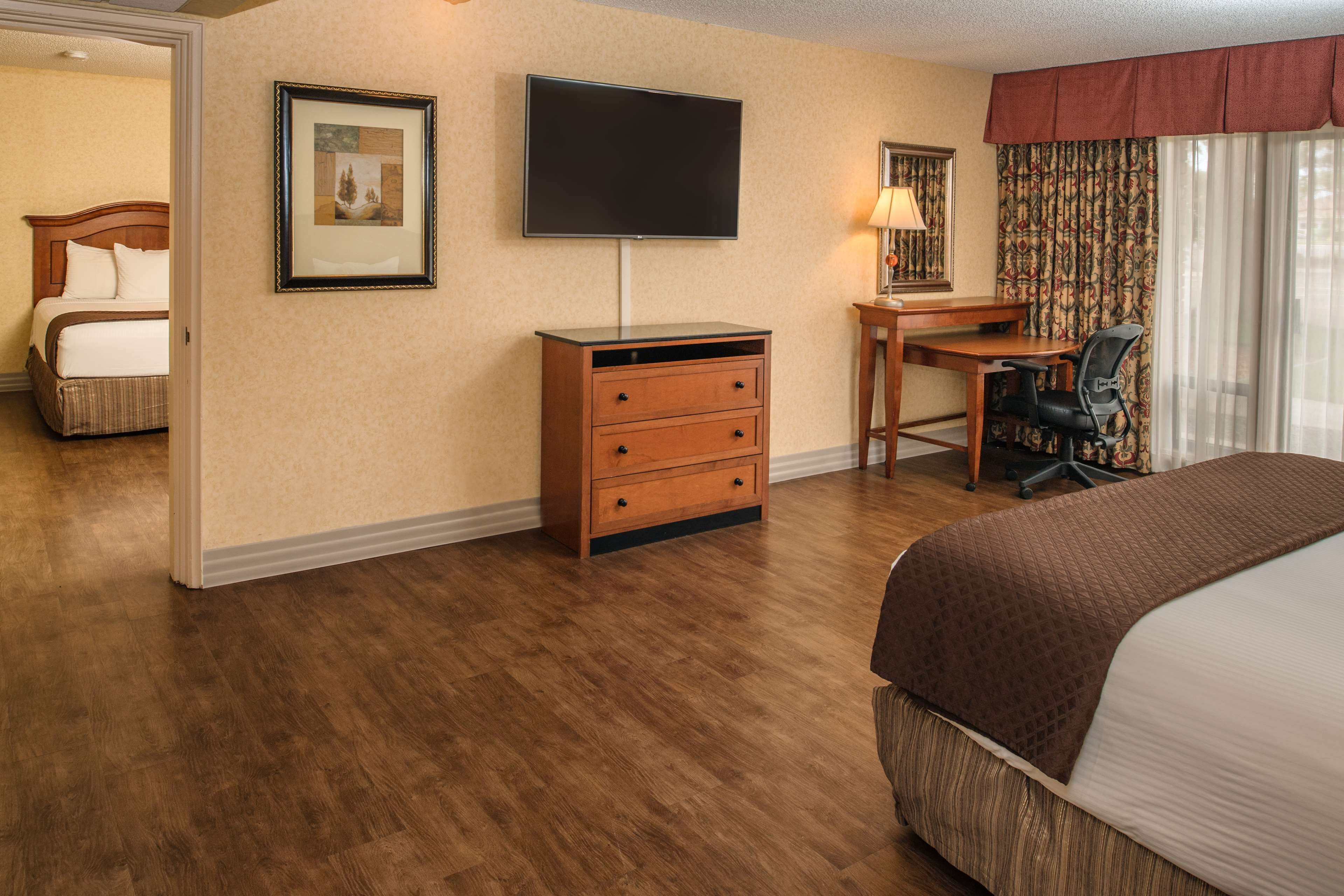 A hotel room with a bed, a desk, a chair, a TV, a dresser, and a door leading to another room.