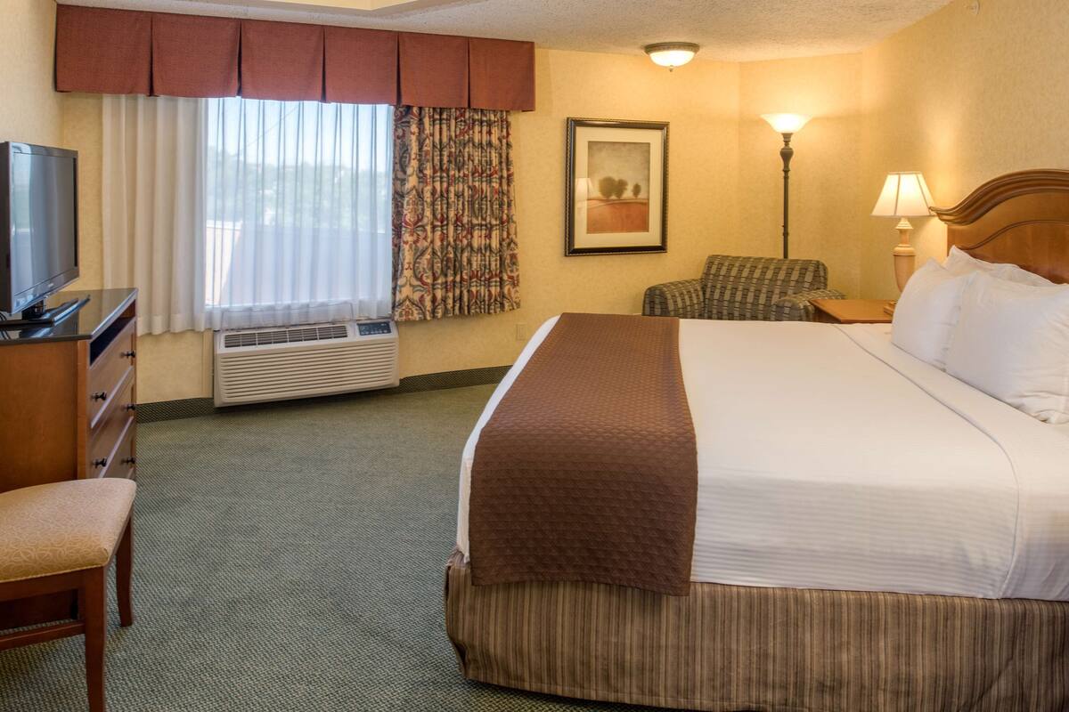 Una habitación de hotel con cama, televisión, una silla, escritorio y lámpara.