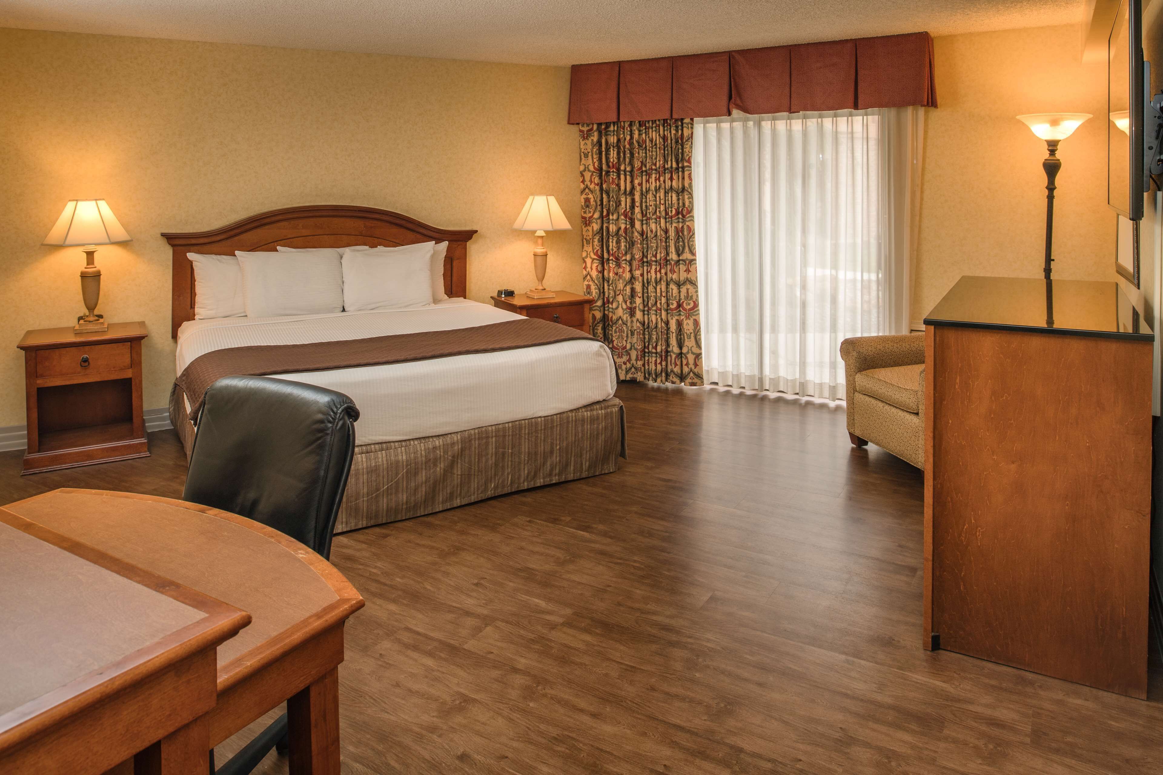 A hotel room with a large bed, two bedside tables with lamps, a wooden desk, a chair, and a large window with curtains.
