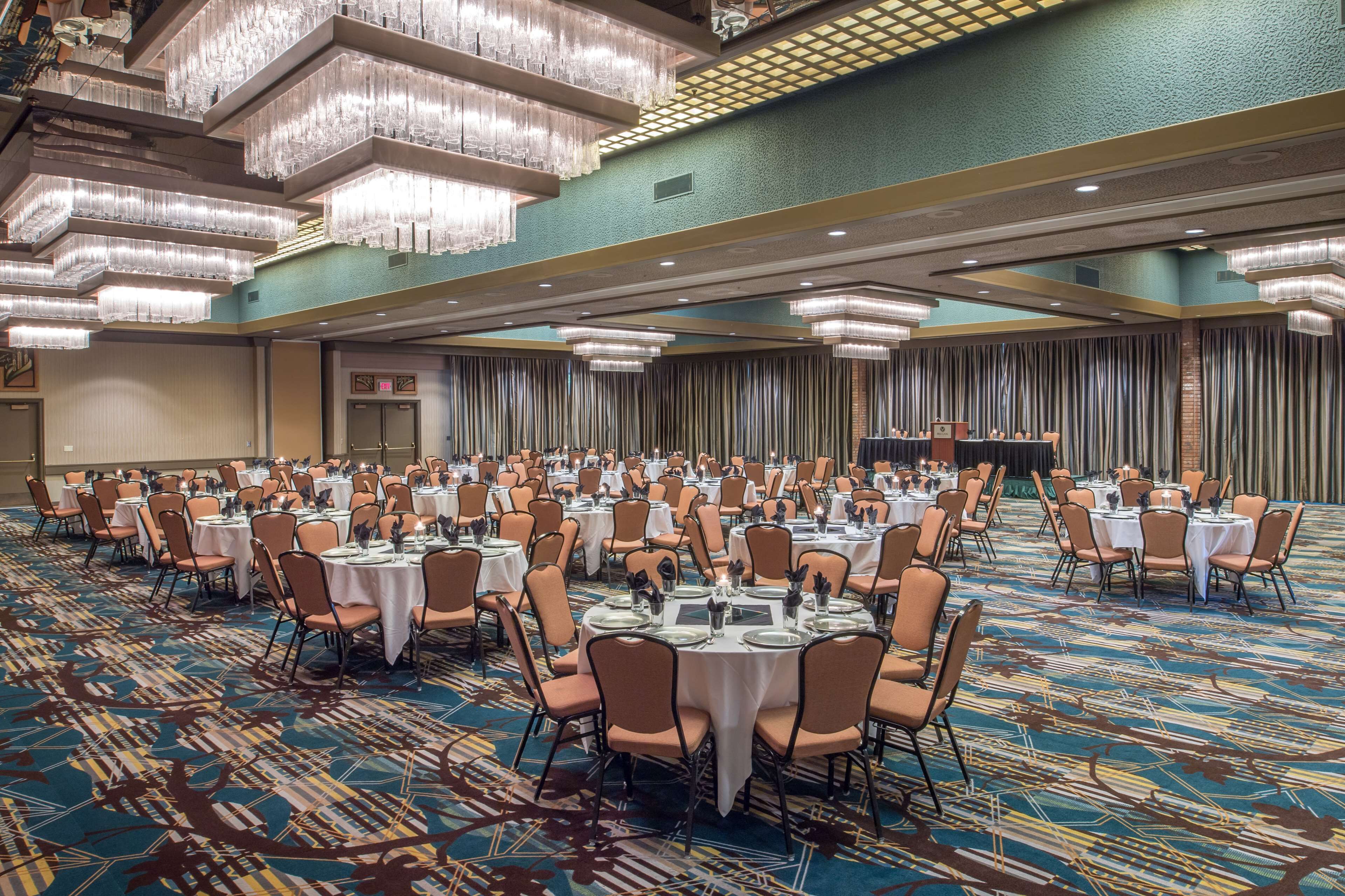 A large banquet hall with round tables and chairs, a patterned carpet, and hanging chandeliers.