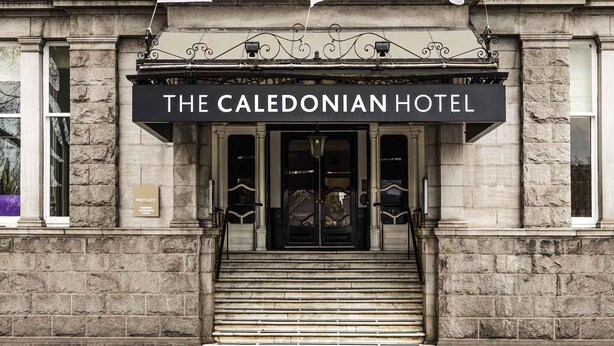 A entrada do Hotel Caledonian com fachada de pedra, uma cobertura de entrada preta e três bandeiras brancas.