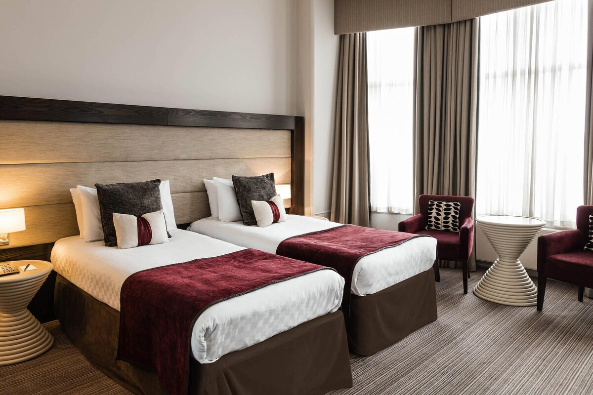 Ein Hotelzimmer mit zwei Betten, einem Sessel, einem Tisch und einem großen Fenster mit Vorhängen.