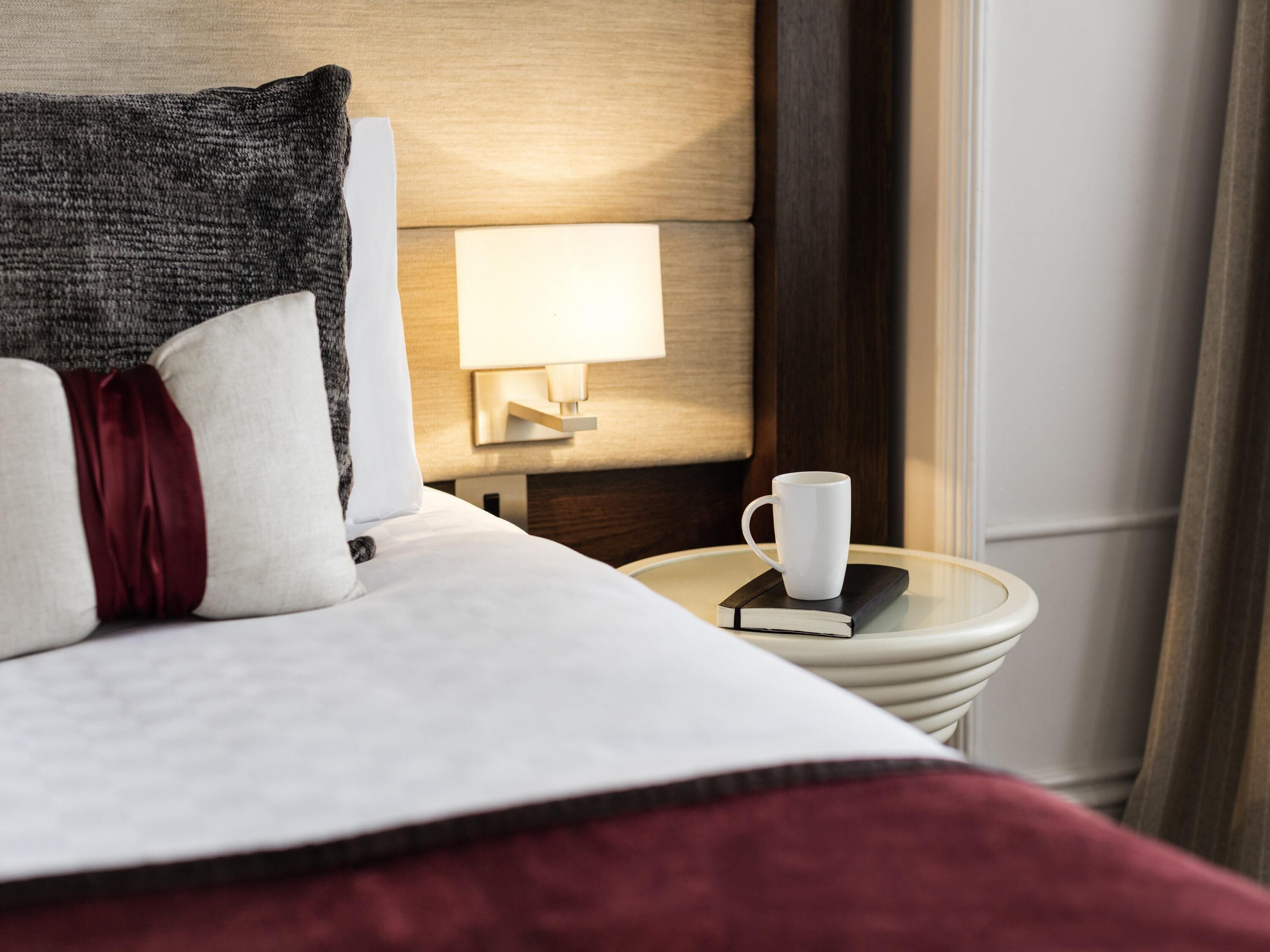 Una cama bien hecha con almohadas, una mesita de noche con un libro y una taza blanca, y una lámpara en la pared.