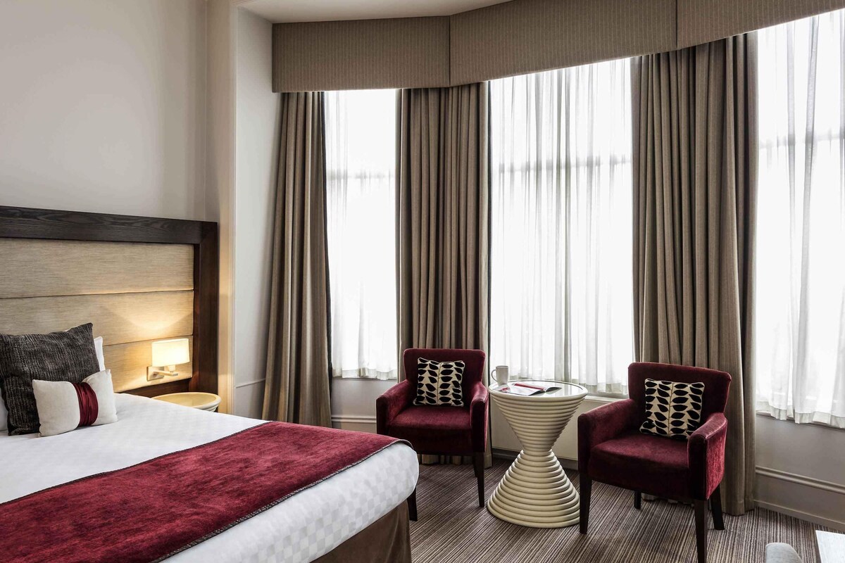 Ein Hotelzimmer mit einem großen Bett, zwei Sesseln, einem kleinen Tisch und einem Fenster mit Vorhängen.