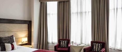 A hotel room with a large bed, two armchairs, a small table, and a window with curtains.