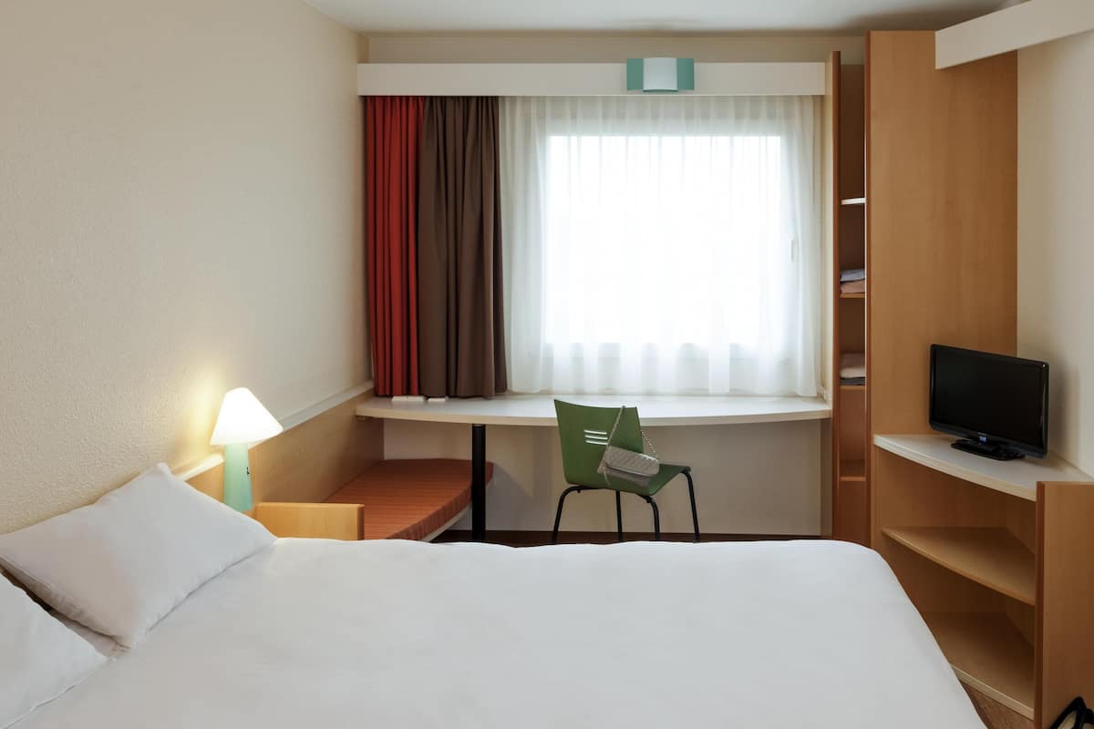 Une chambre d’hôtel avec un lit, un bureau et une chaise, une télévision et une fenêtre avec des rideaux.