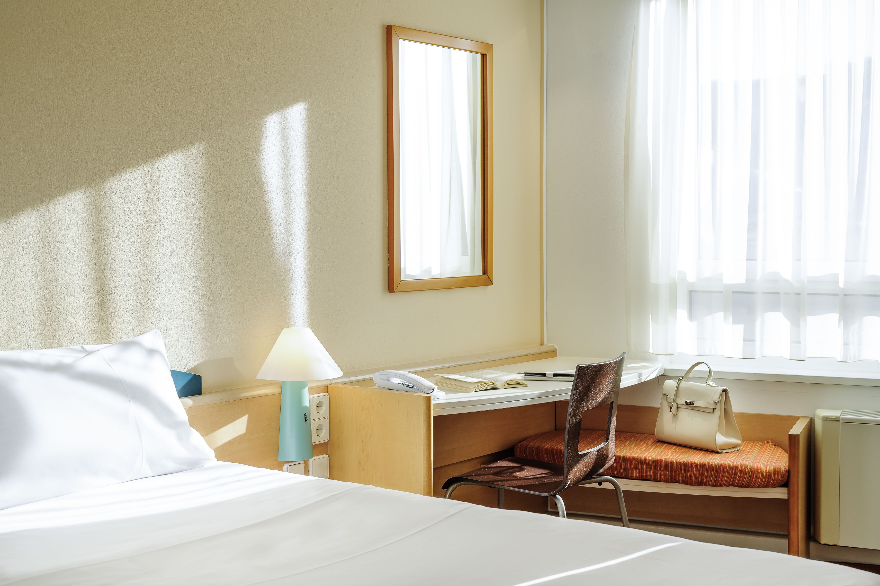 Ein Hotelzimmer mit Bett, Schreibtisch mit Stuhl, Lampe, Handtasche und einem Fenster mit Vorhängen.