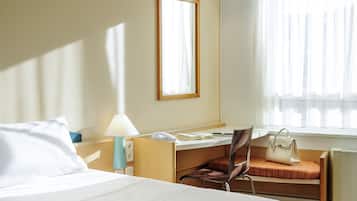 Ein Hotelzimmer mit Bett, Schreibtisch mit Stuhl, Lampe, Handtasche und einem Fenster mit Vorhängen.