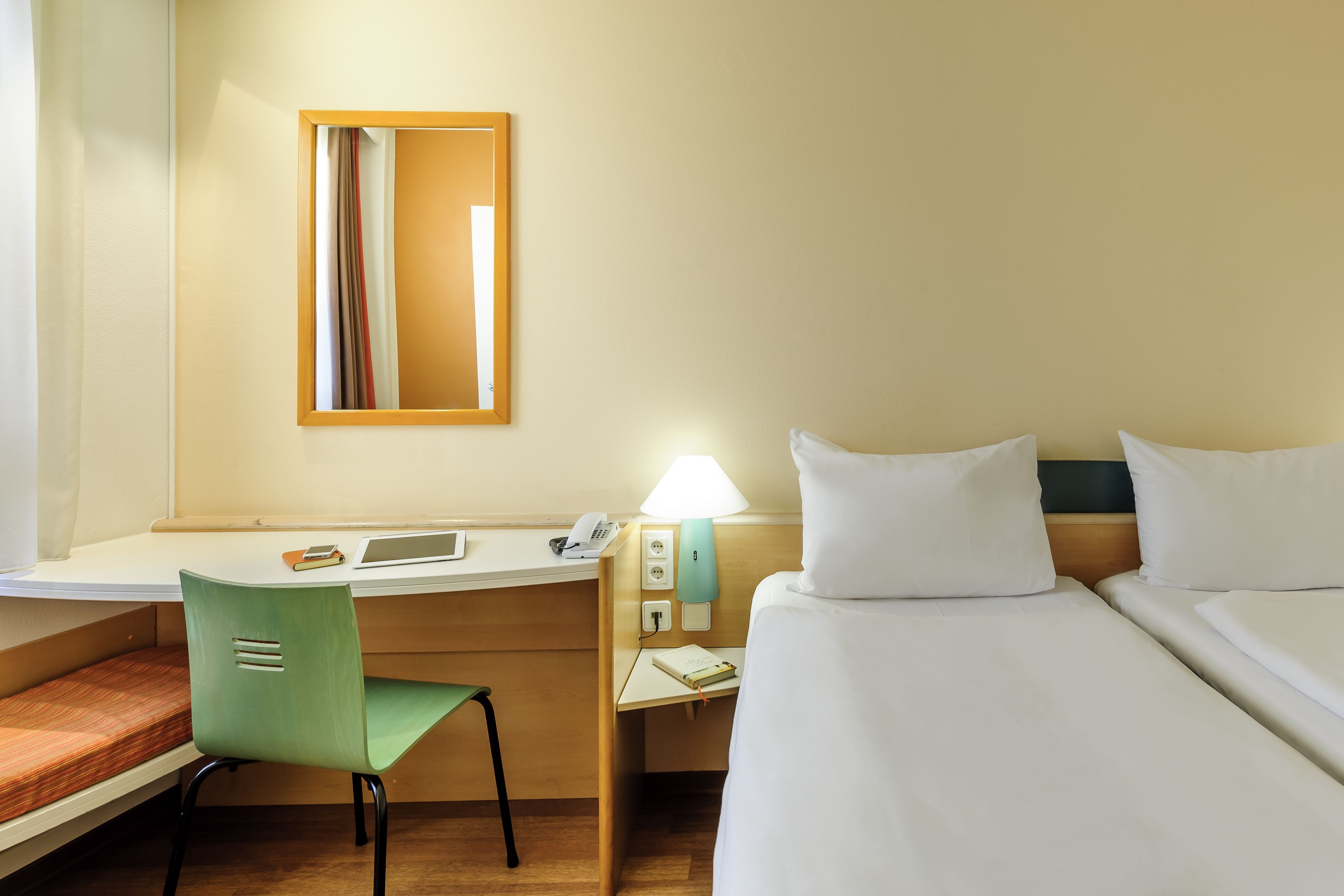 Habitación de hotel con escritorio, una silla verde, cama con ropa de cama blanca, mesita de noche con lámpara y un espejo en la pared.
