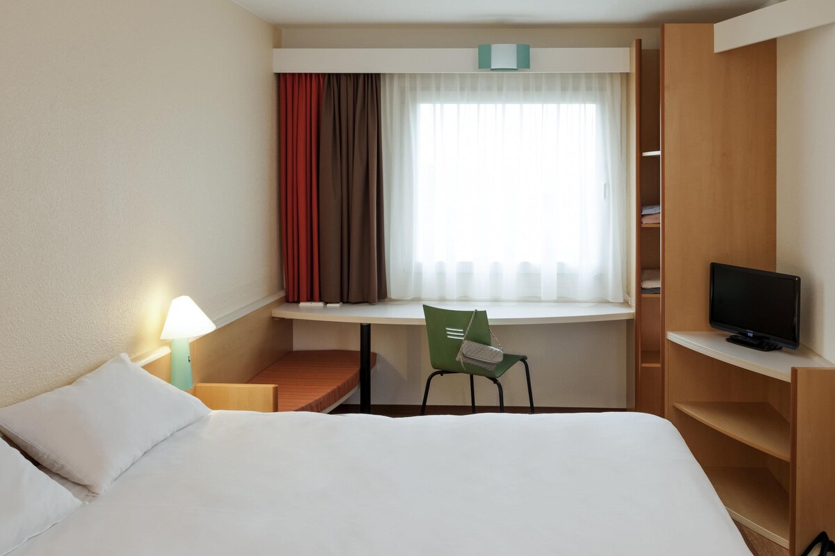 Une chambre d’hôtel avec un lit, un bureau et une chaise, une télévision et une fenêtre avec des rideaux.