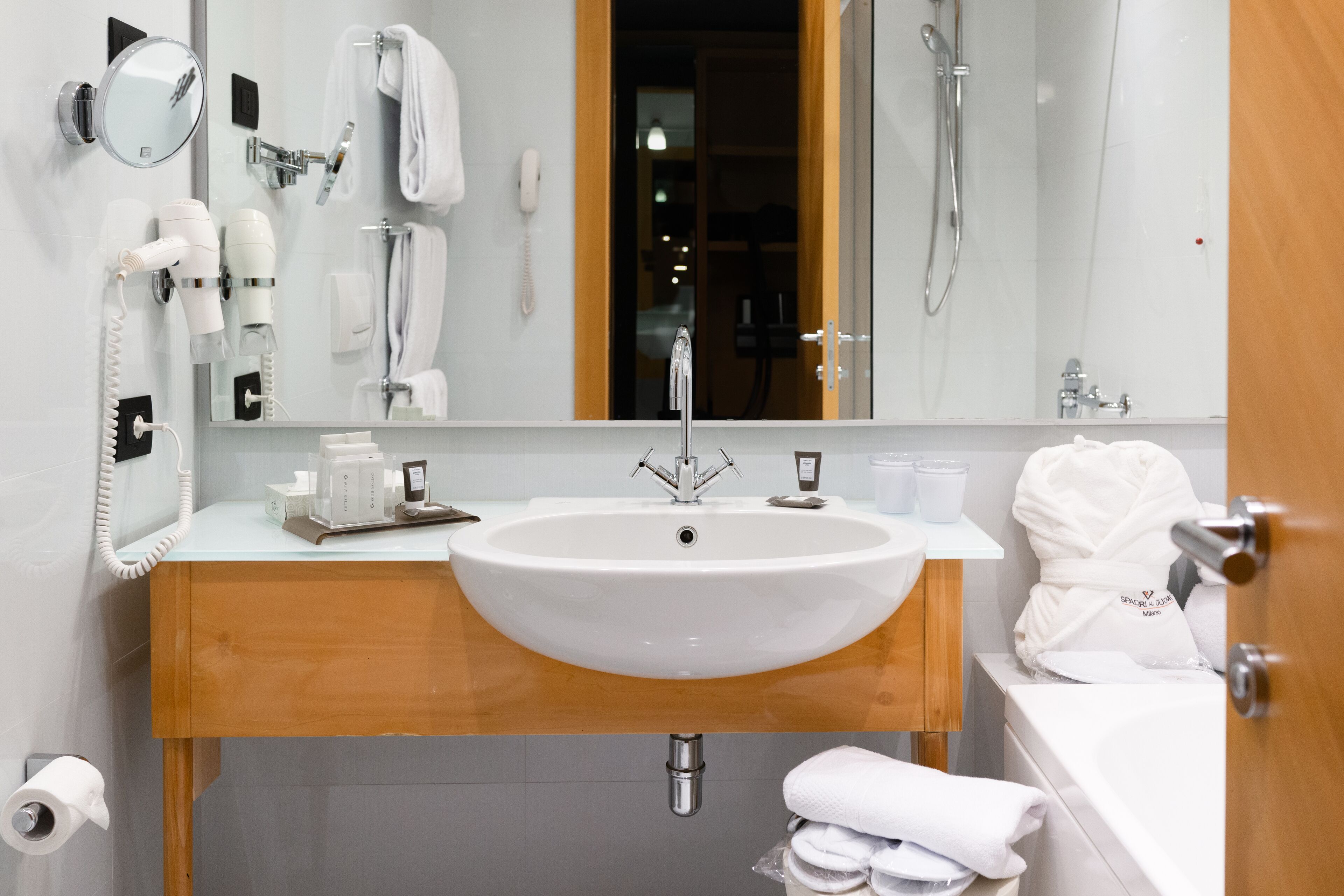 A modern bathroom with a large sink, a mirror, a hairdryer, a telephone, and a shower.