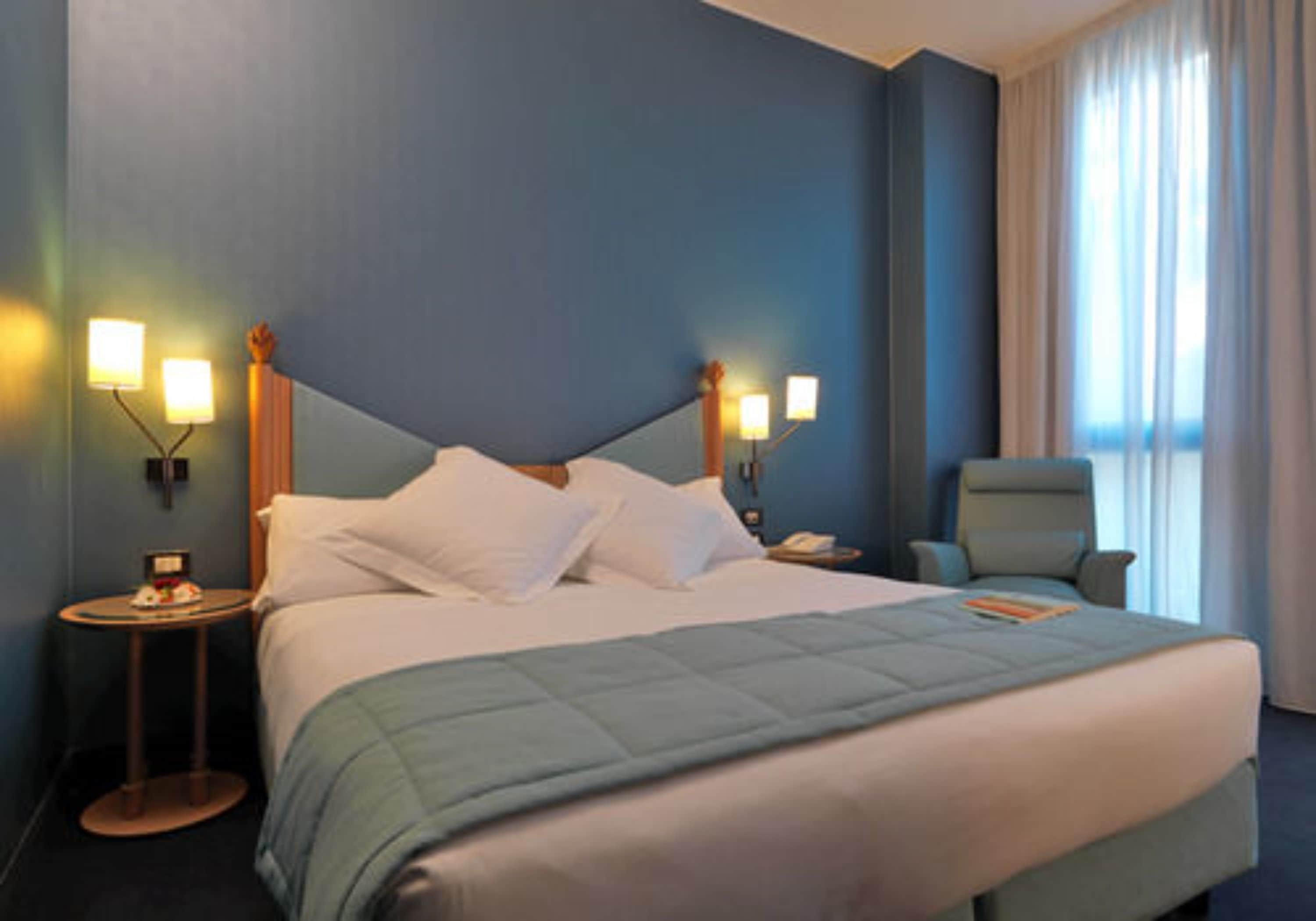 Una habitación de hotel con una cama grande, dos mesitas de noche con lámparas, una silla y un ventanal con cortinas.