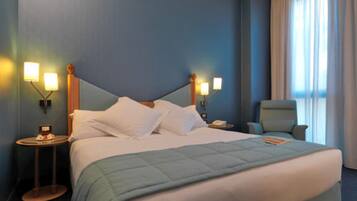 Una habitación de hotel con una cama grande, dos mesitas de noche con lámparas, una silla y un ventanal con cortinas.