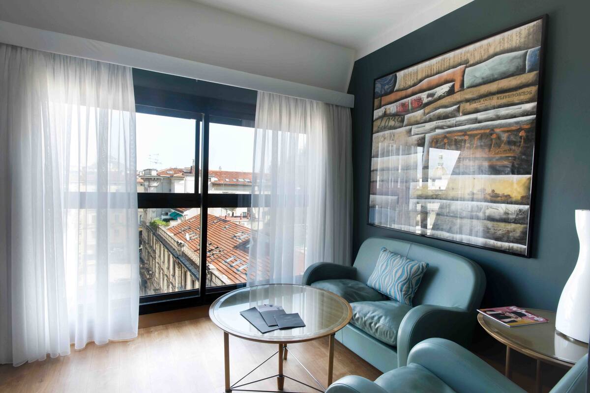 A modern living room with a large window, a round glass coffee table, a blue sofa, and a large abstract painting on the wall.