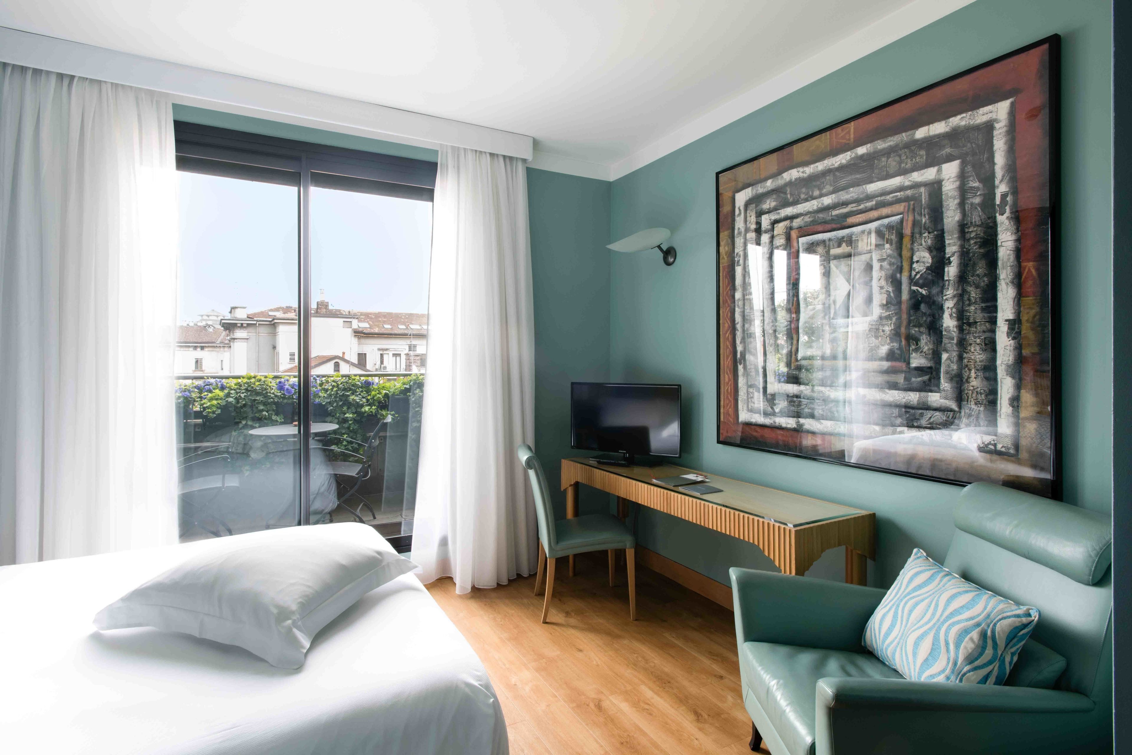 A modern hotel room with a bed, a desk with a TV, a green sofa, and a large window offering a city view.