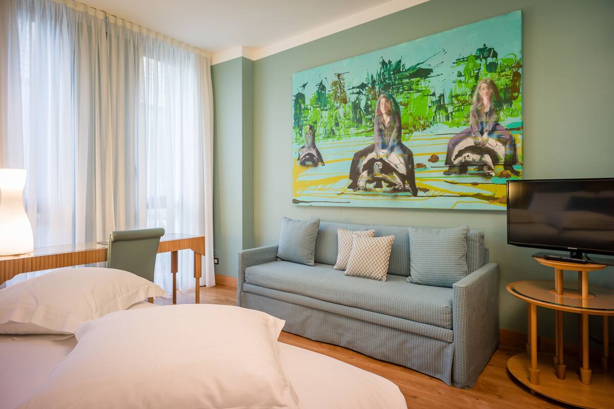 A hotel room with a bed, a sofa, a TV, and a painting on the wall.
