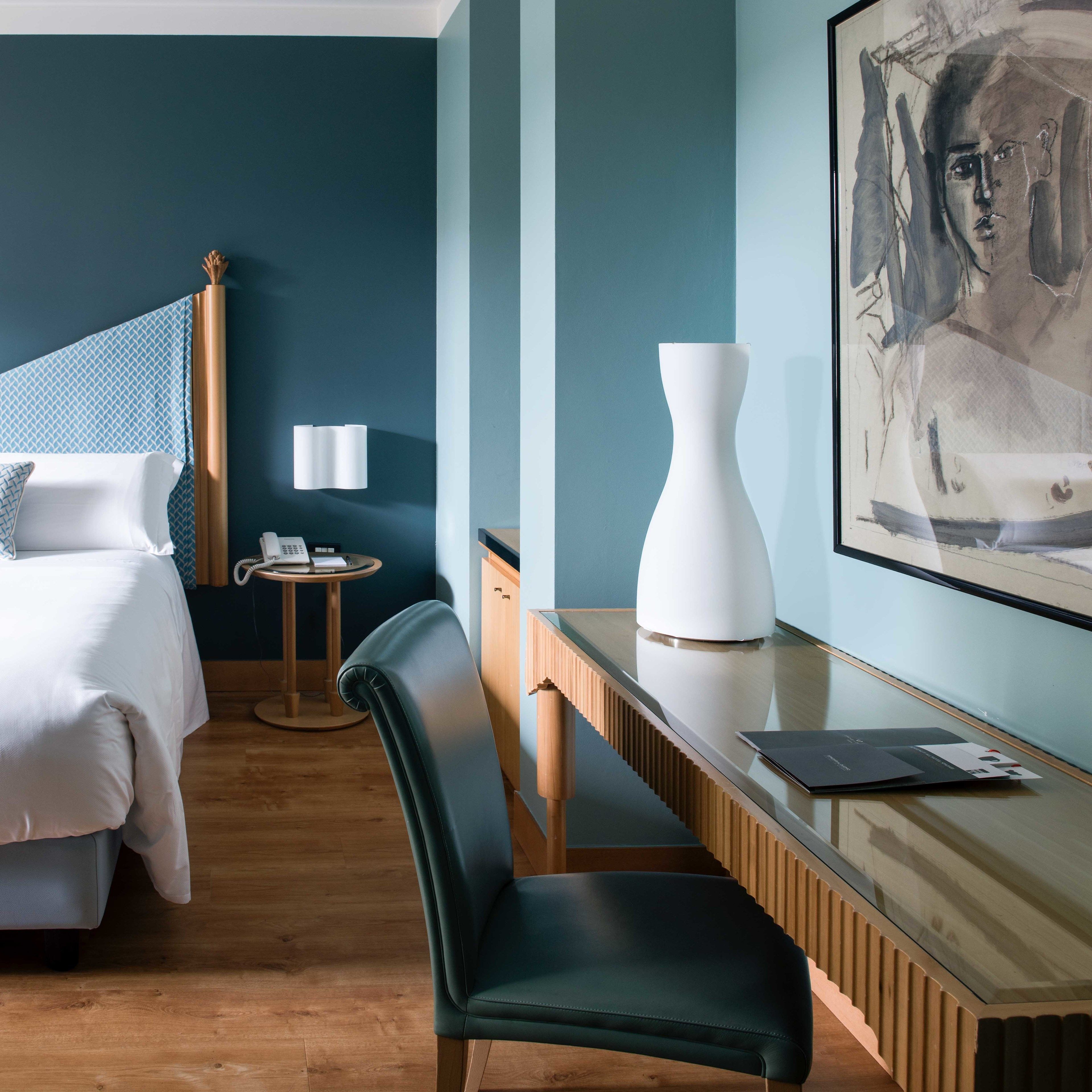 Een hotelkamer met een bed, een bureau, een stoel, een lamp en een schilderij aan de muur.