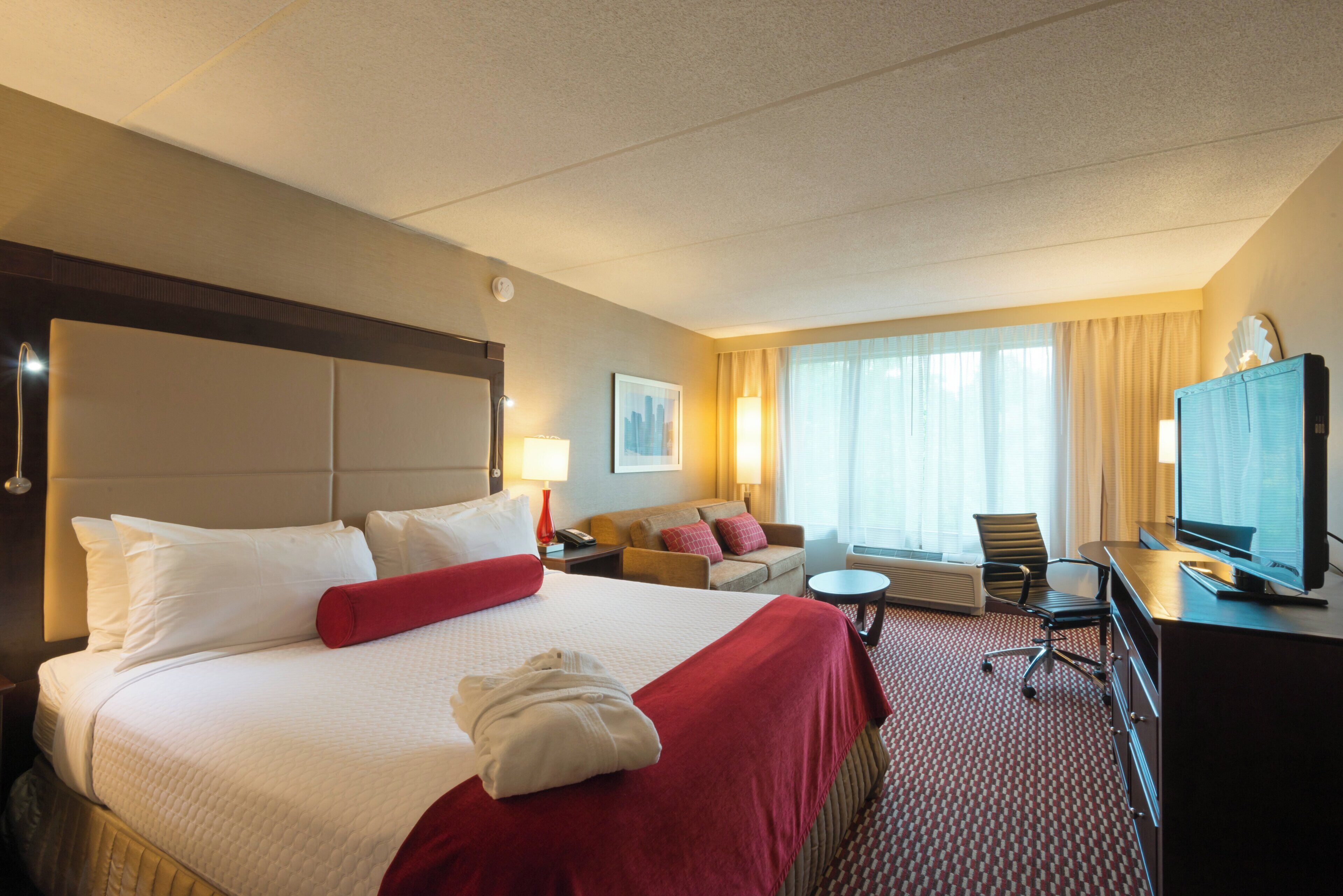 A hotel room with a large bed, a sofa, a desk with a television, and a window with curtains.