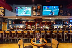 Un bar con varias pantallas mostrando eventos deportivos, una variedad de botellas y una zona de estar con mesas y sillas.