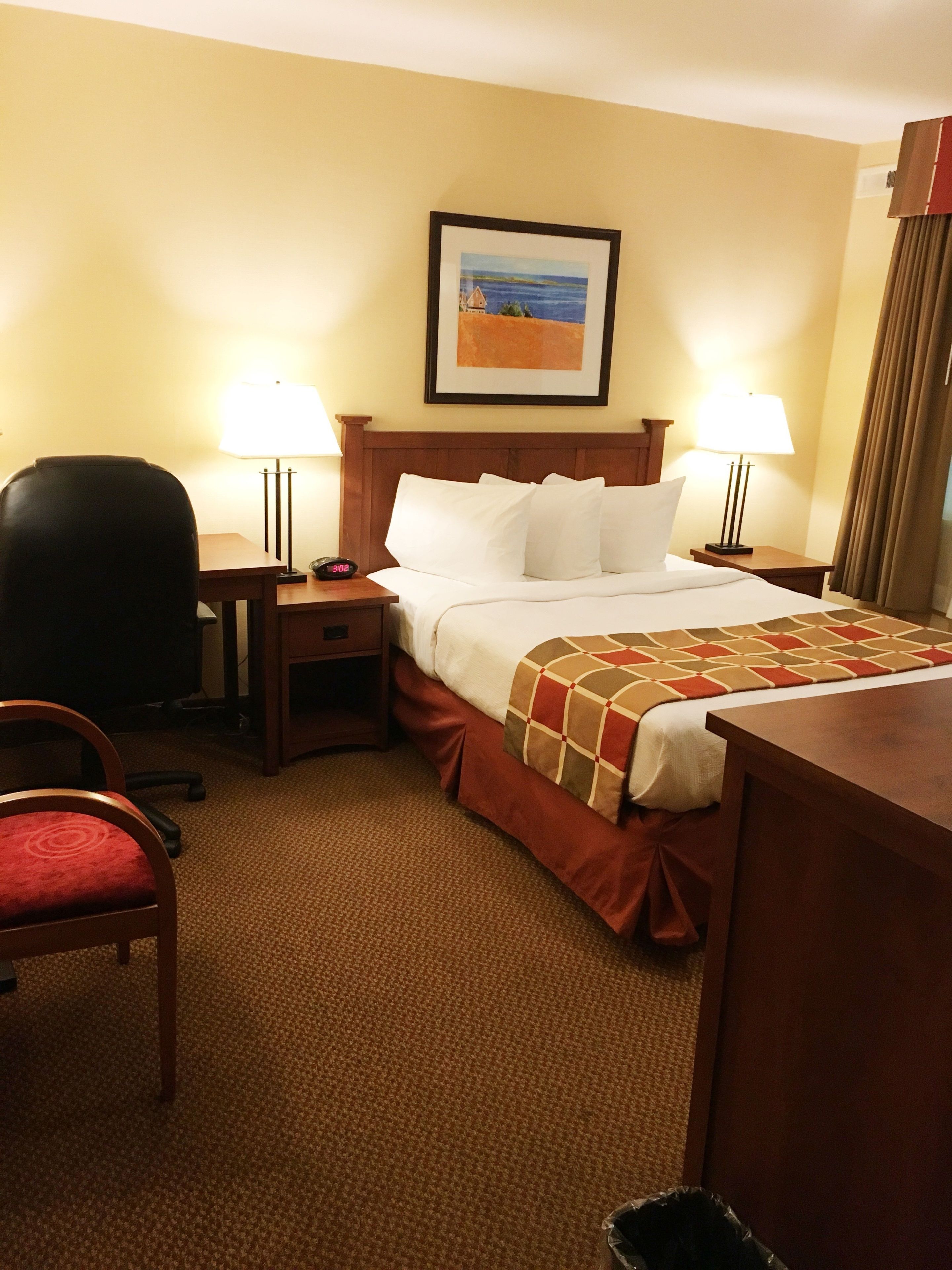 Habitación de hotel con una cama, dos lámparas de noche, una silla, un escritorio y un cuadro en la pared.