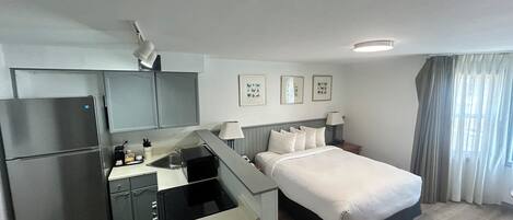 Una habitación de hotel compacta con kitchenette, una cama, un escritorio y una ventana con cortinas.
