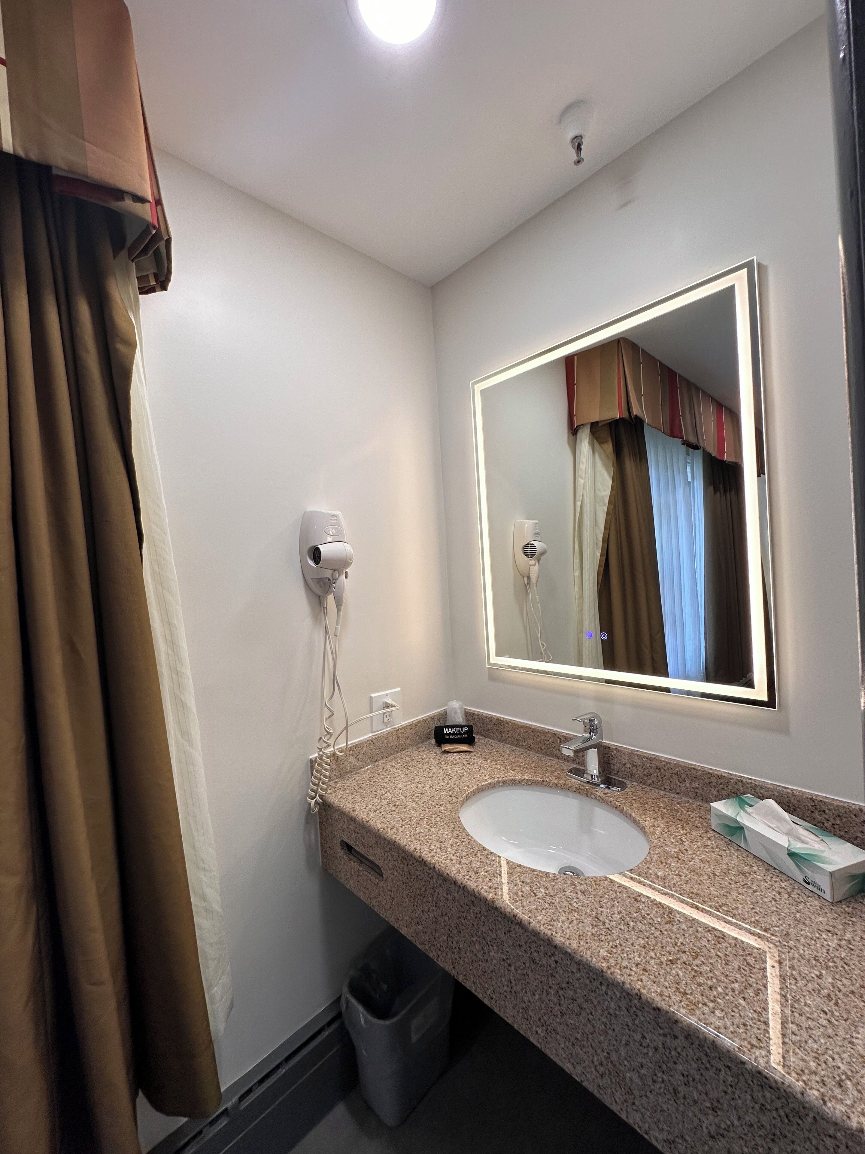 A hotel bathroom with a granite countertop sink, a large mirror, a hairdryer, and a trash bin.