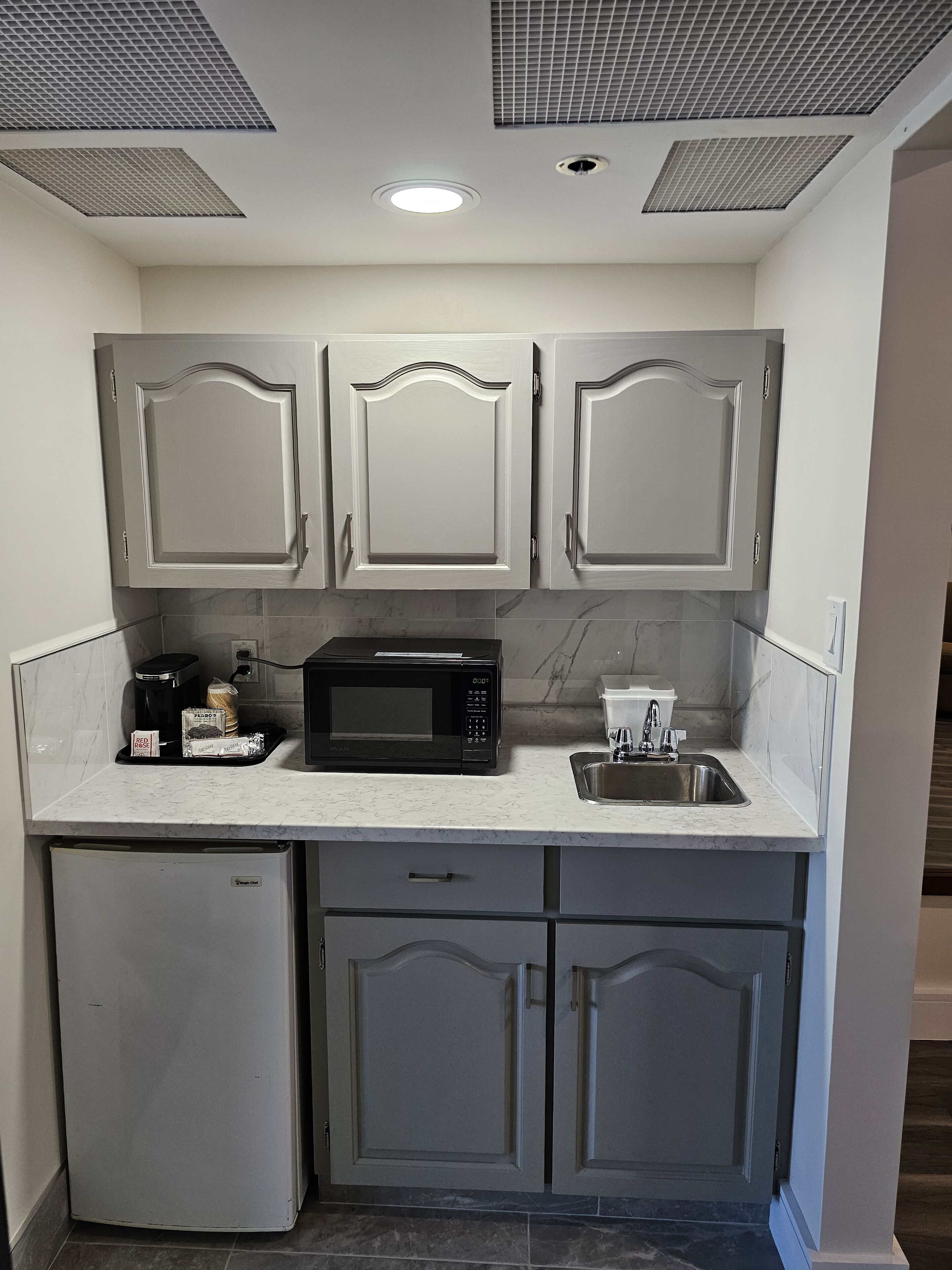 Una cocina compacta con armarios blancos, un microondas negro, una cafetera y un refrigerador pequeño.