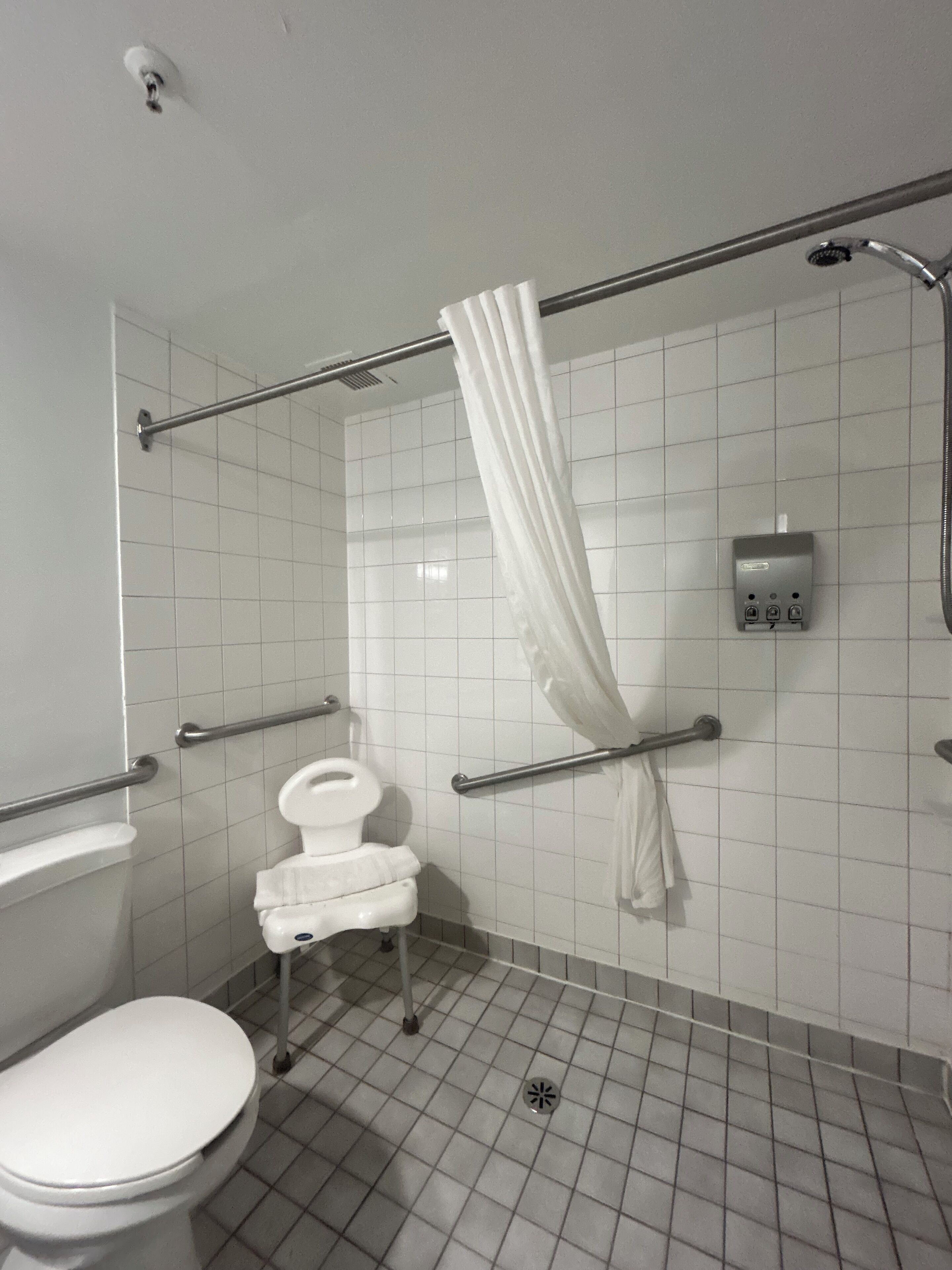 Une salle de bain avec des toilettes blanches, une barre d’appui, une douche avec un rideau blanc et un sèche-mains.