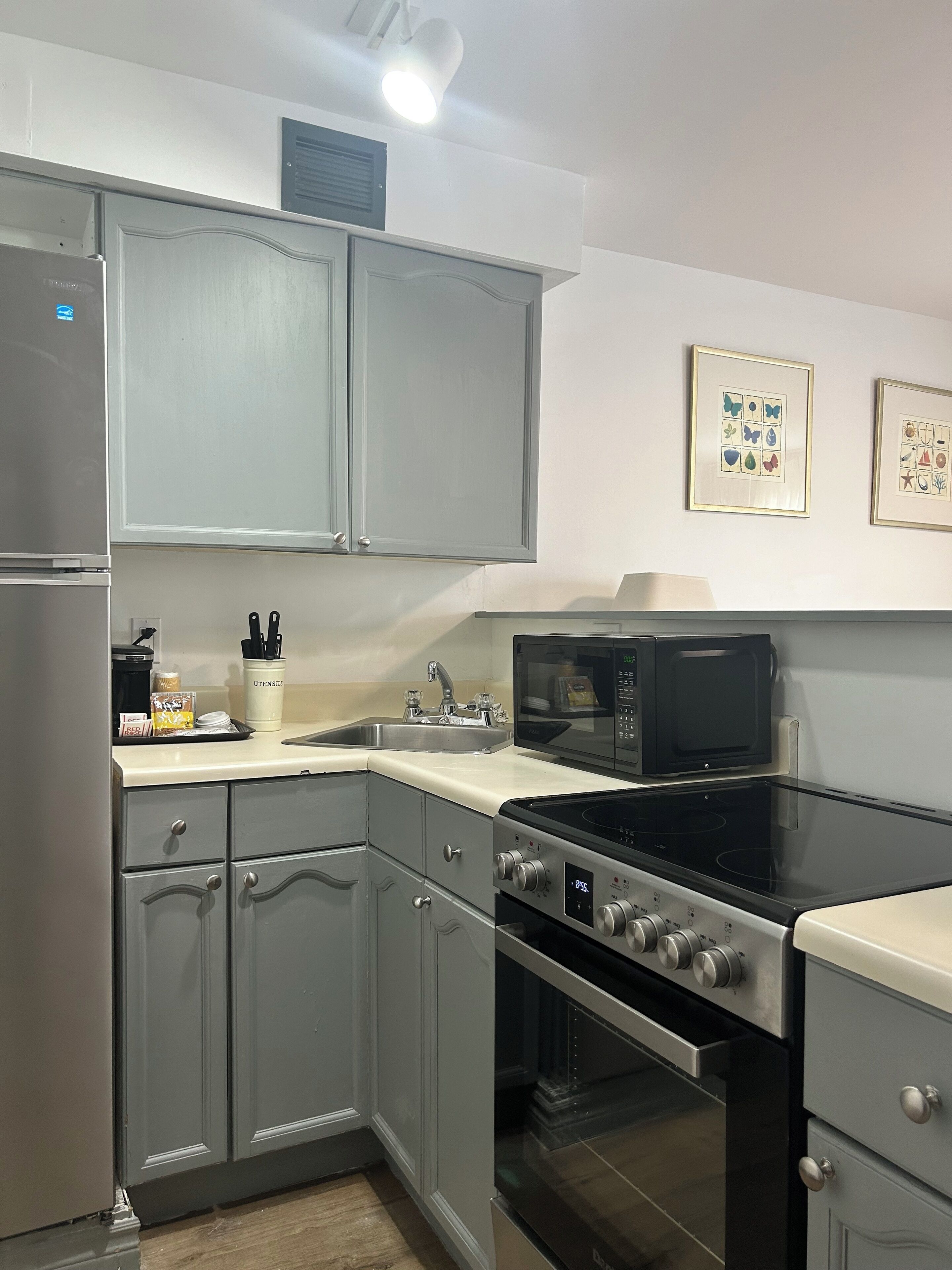 A kitchen with grey cabinets, a stainless steel refrigerator, a microwave, an oven, and a sink. There are framed pictures on the wall and a coffee maker on the counter.