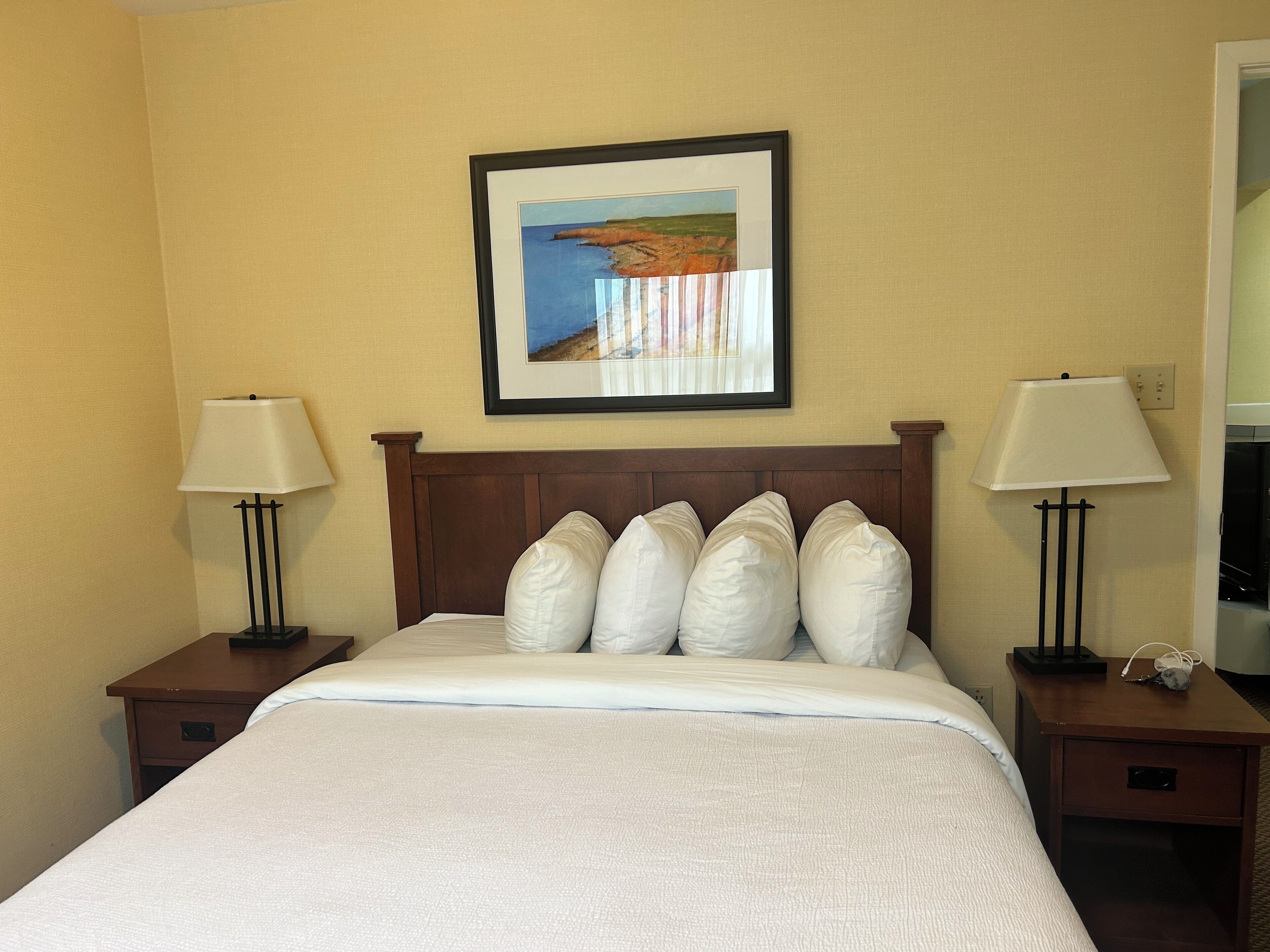 A hotel room with a bed, two bedside tables, two lamps, a framed picture on the wall, and a nightstand with a phone.