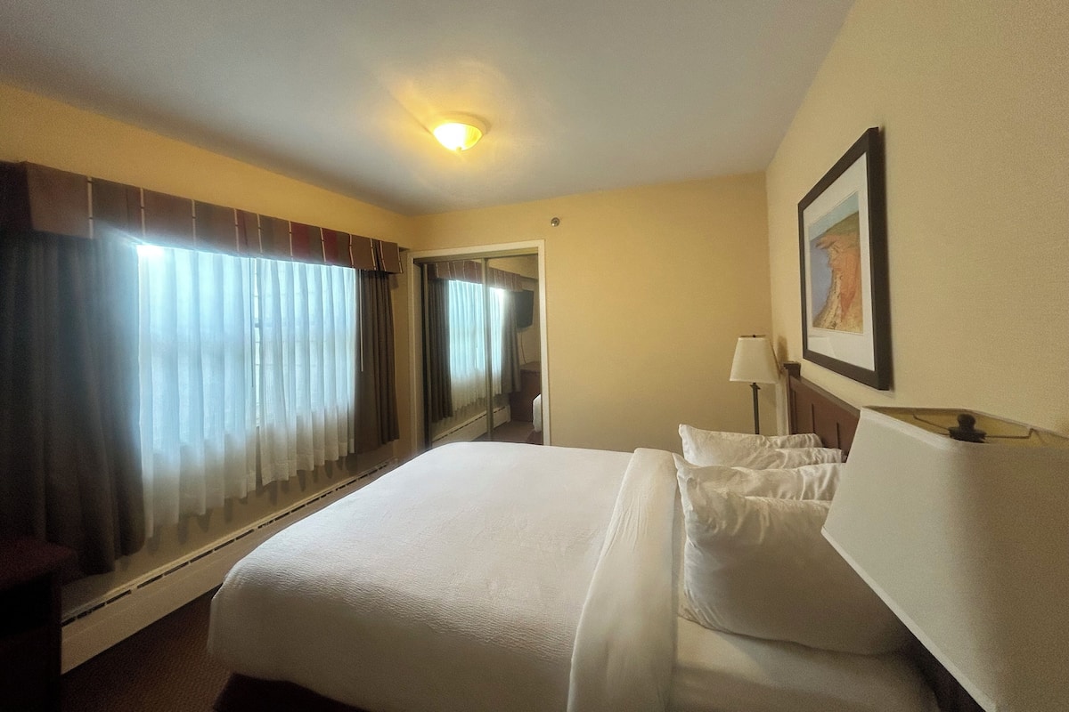 A hotel room with a large bed, a bedside table with a lamp, a framed picture on the wall, and a window with curtains.