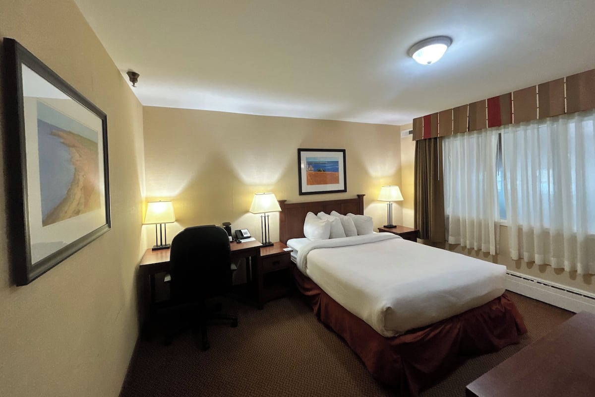 A hotel room with a bed, two bedside tables with lamps, a desk with a chair, a framed picture on the wall, and a window with curtains.