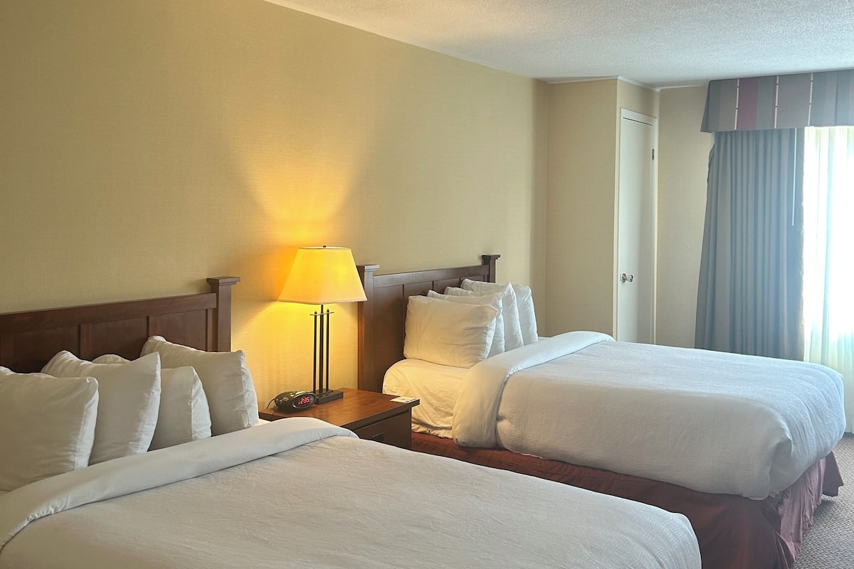 A hotel room with two beds, a nightstand with a lamp, and a window with curtains.