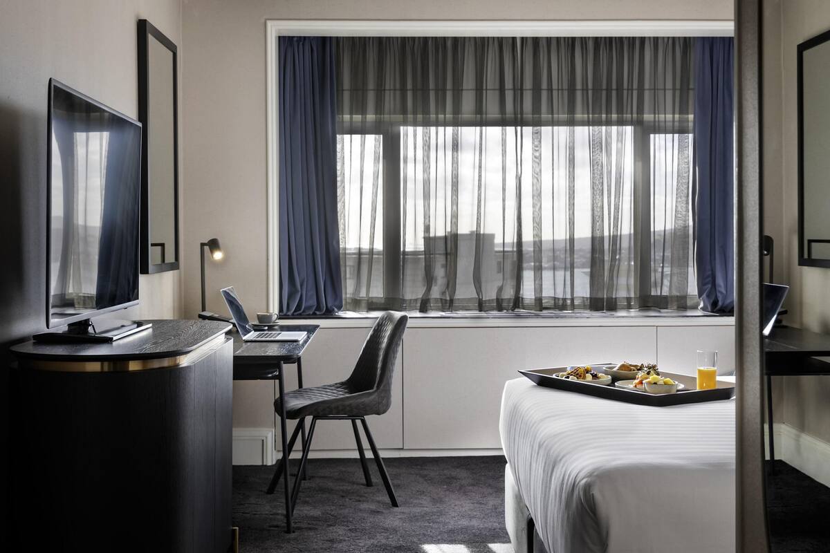 A hotel room with a bed, a desk with a laptop, a chair, a TV, and a window with curtains.