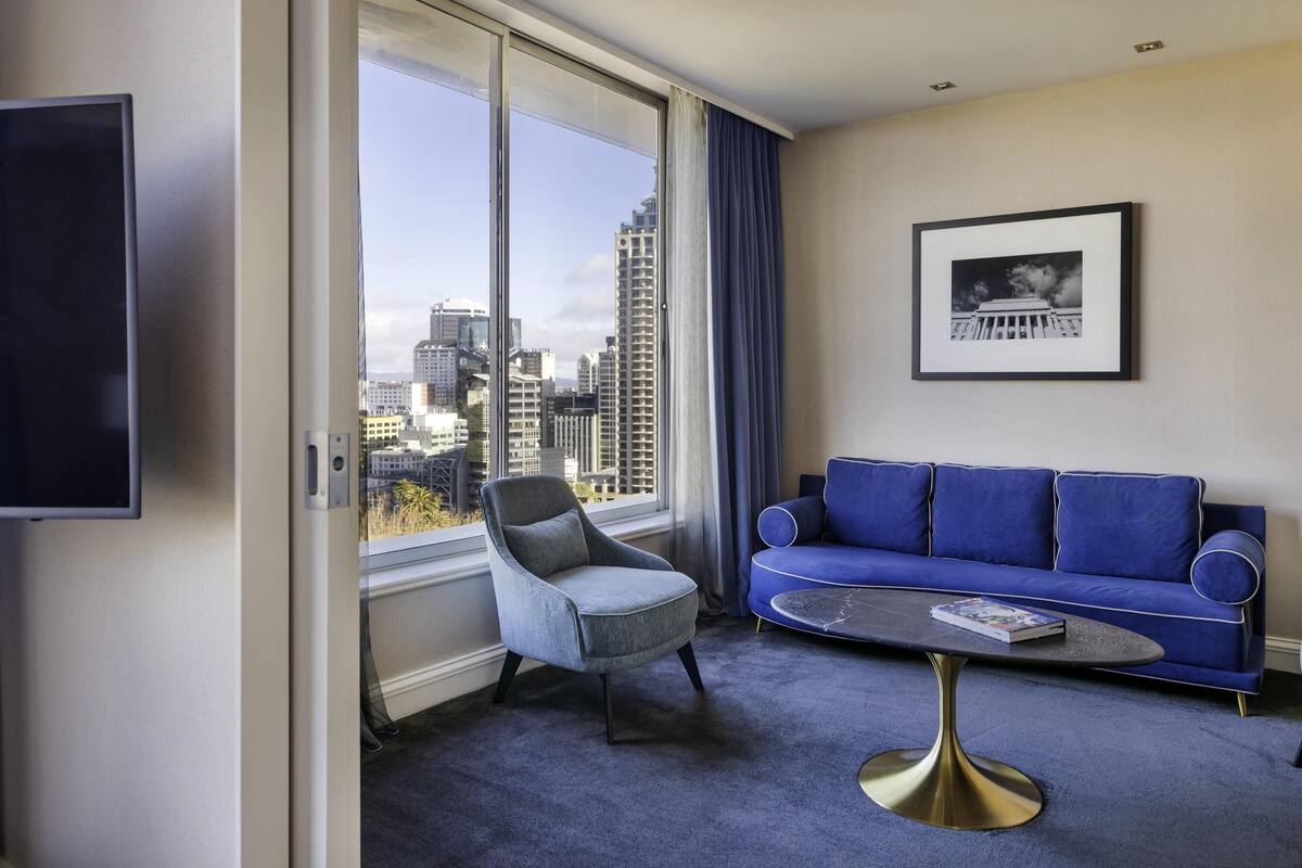 A modern hotel room with a large window, a blue sofa, a grey armchair, a round coffee table, and a framed picture on the wall.