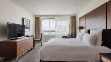 A hotel room with a large bed, a TV, a desk, and a view of the city through the window.