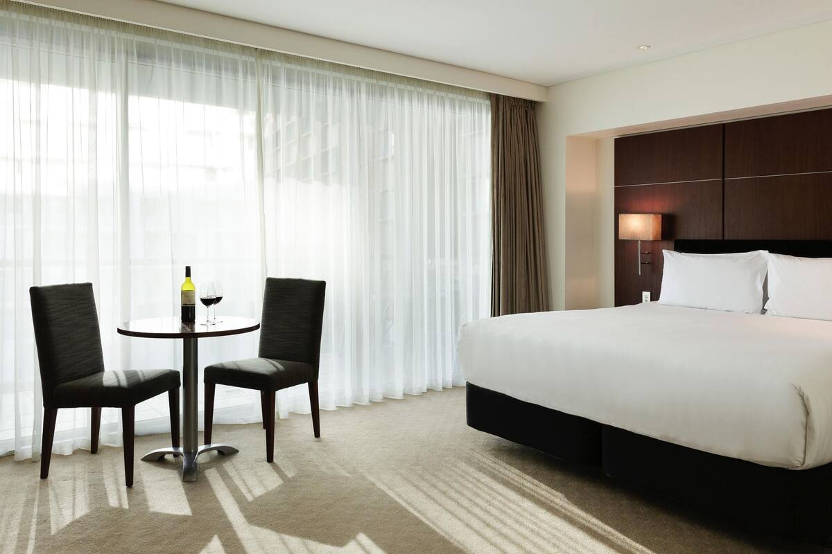 A hotel room with a bed, two chairs, a small table with a bottle and glass, and sheer curtains.