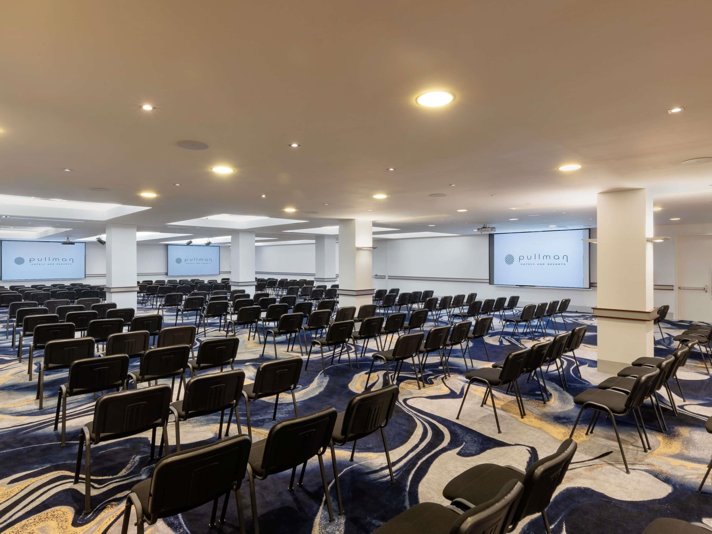 A conference room with rows of chairs, a large screen, and a carpeted floor.