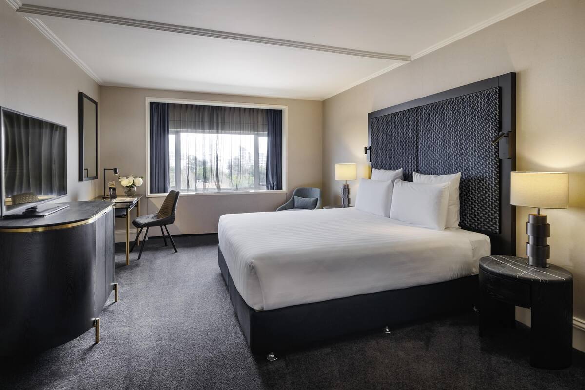 A hotel room with a large bed, a desk, a chair, a nightstand, and a window with curtains.