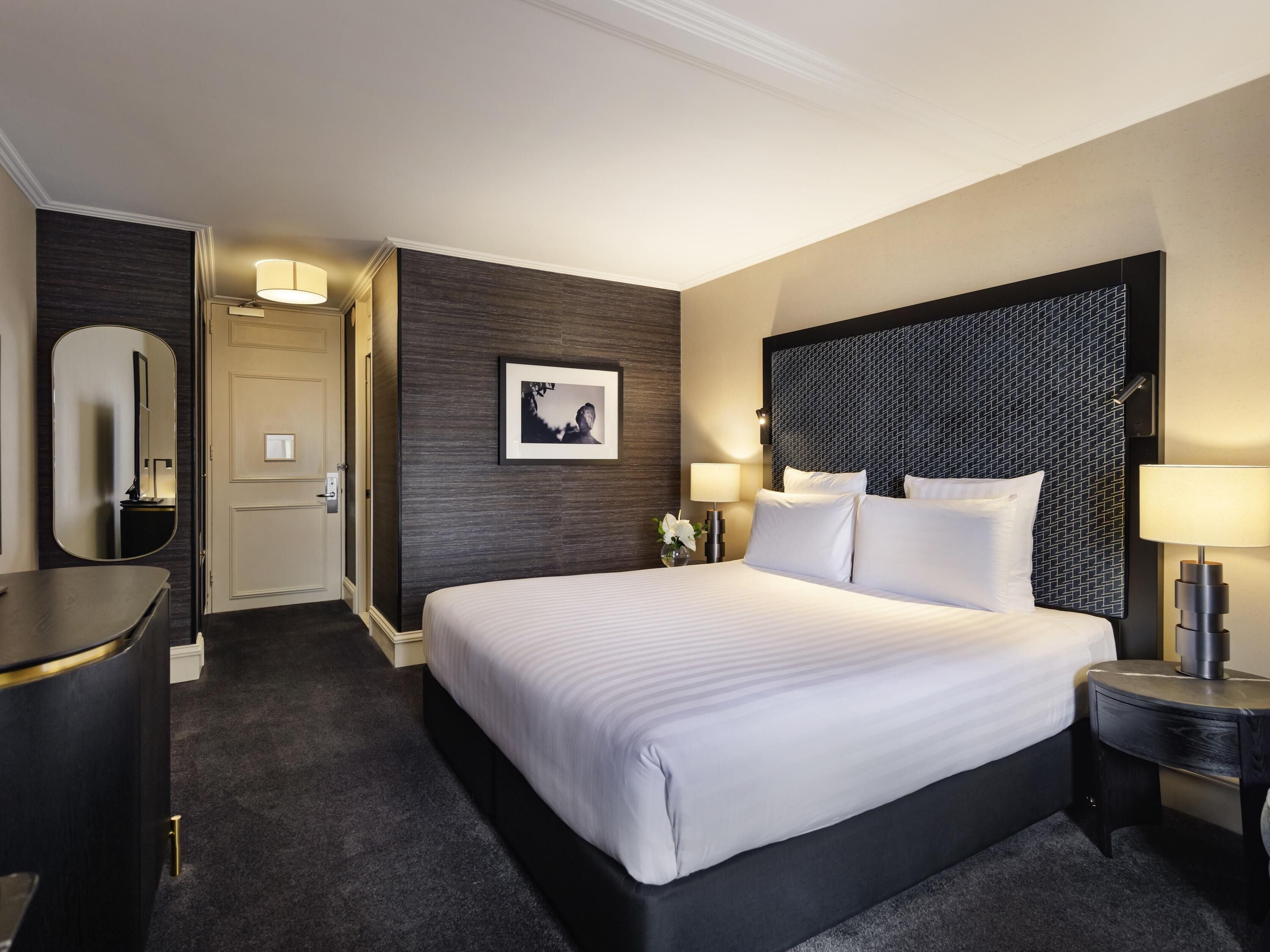 A hotel room with a large bed, a desk, a chair, a mirror, and a picture on the wall.