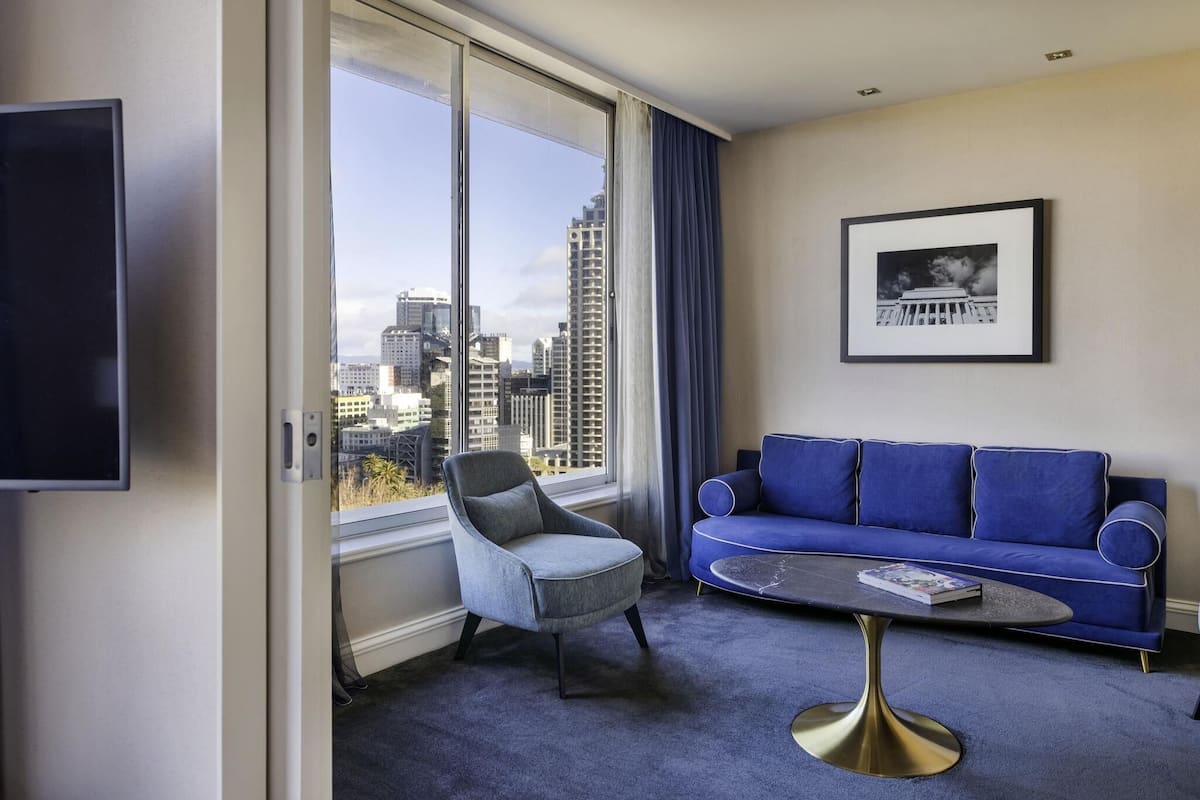 A modern hotel room with a large window, a blue sofa, a grey armchair, and a round coffee table.