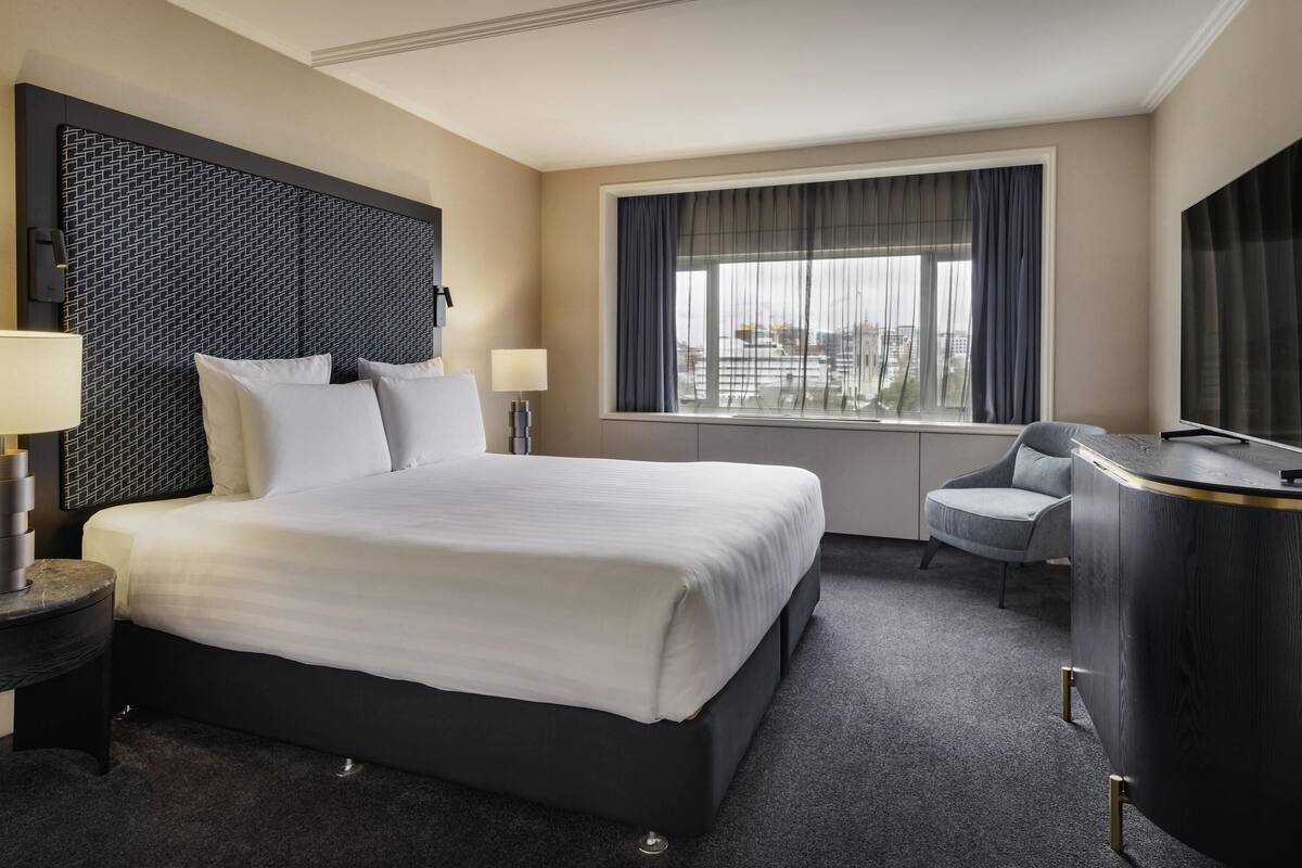 A hotel room with a large bed, a desk, a chair, a TV, and a window with curtains.