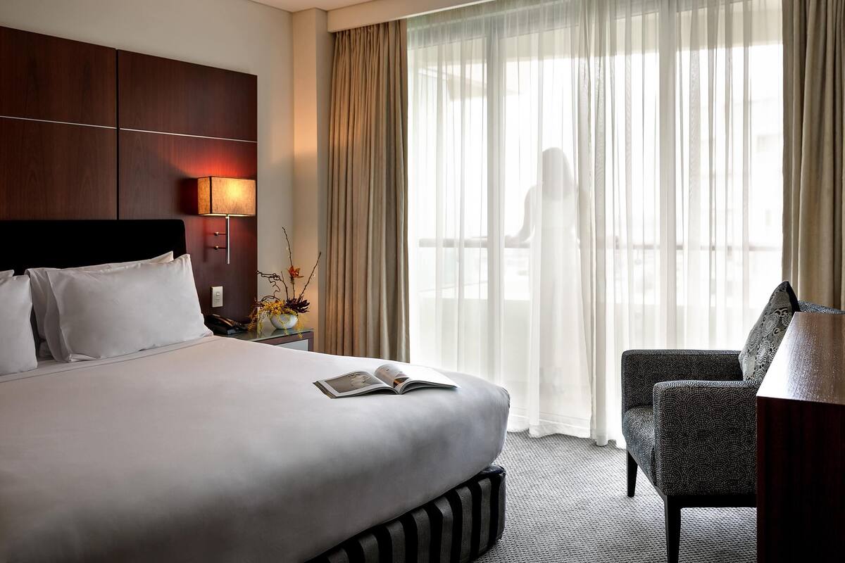 A hotel room with a large bed, a bedside table with a lamp, a chair, and a view of a balcony.