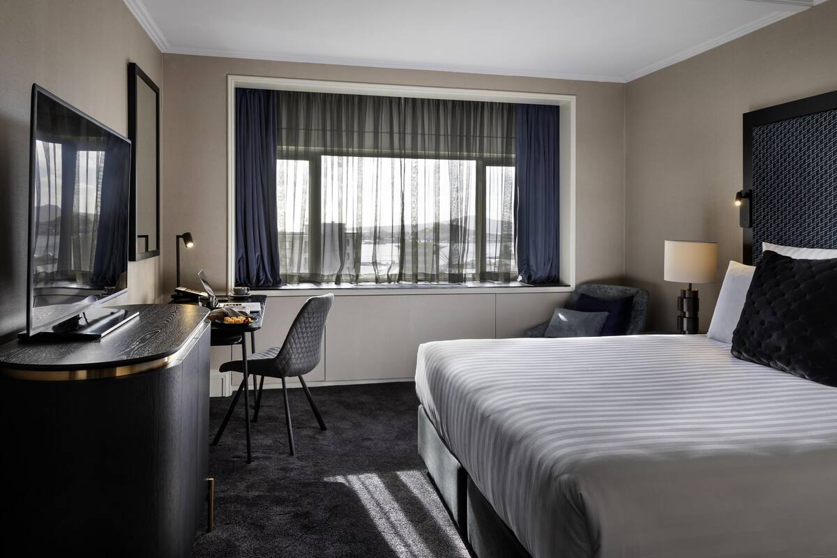 Habitación de hotel con cama, escritorio, silla, televisor y ventana con cortinas.