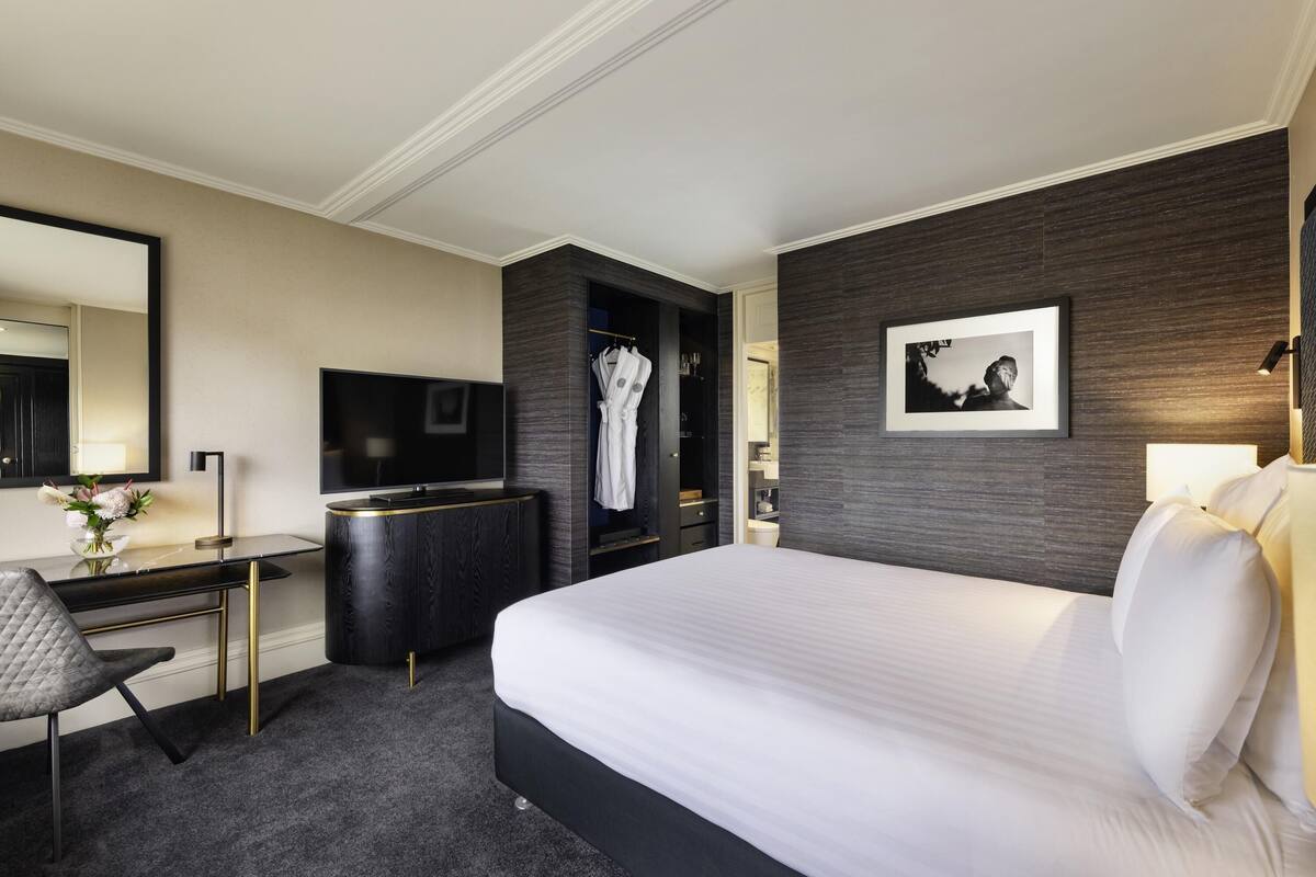 A hotel room with a large bed, a desk with a chair, a TV, a robe hanging, and a framed picture on the wall.