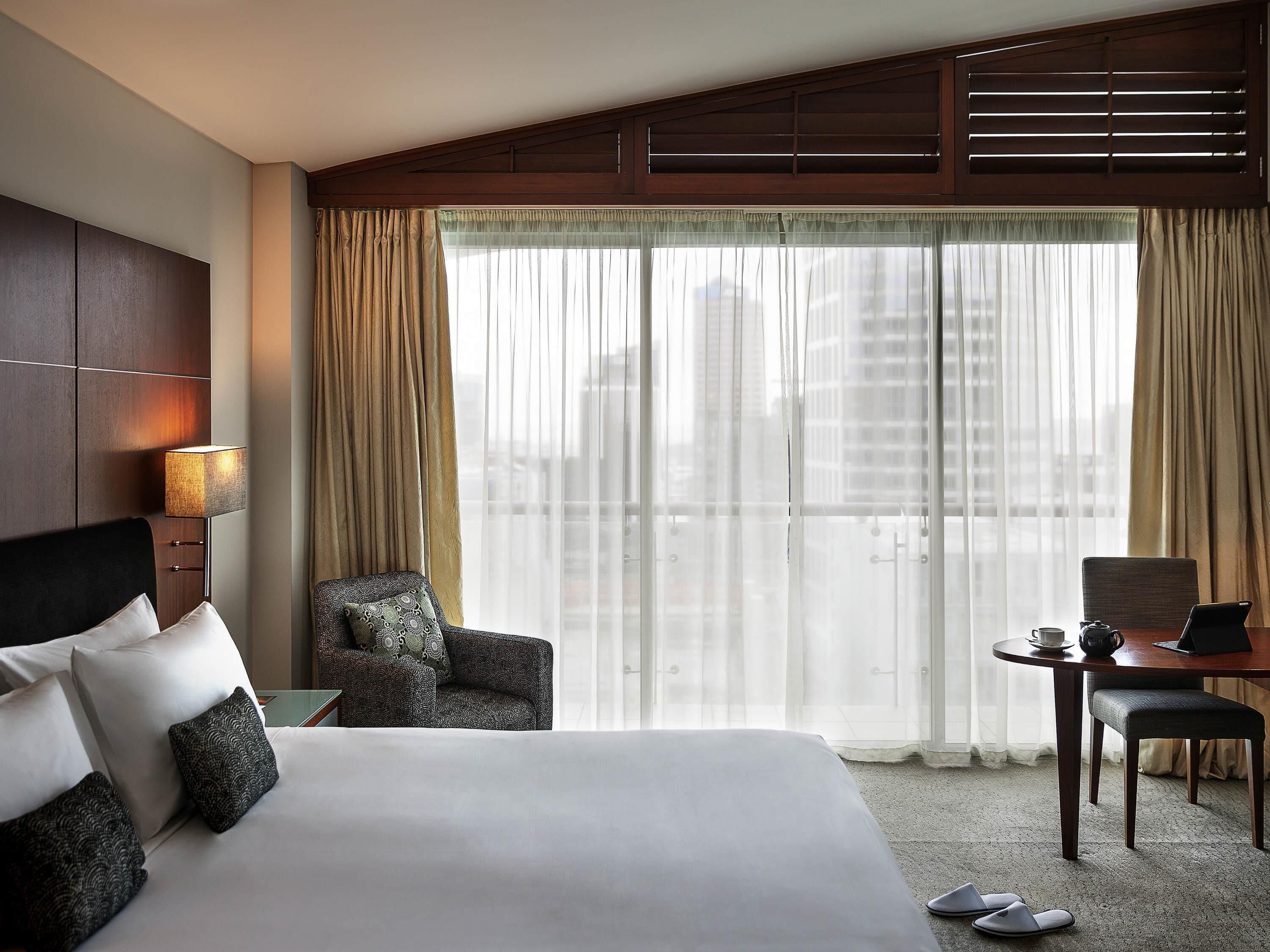 Habitación de hotel con una cama grande, un escritorio, una silla y vistas a la ciudad a través de cortinas translúcidas.