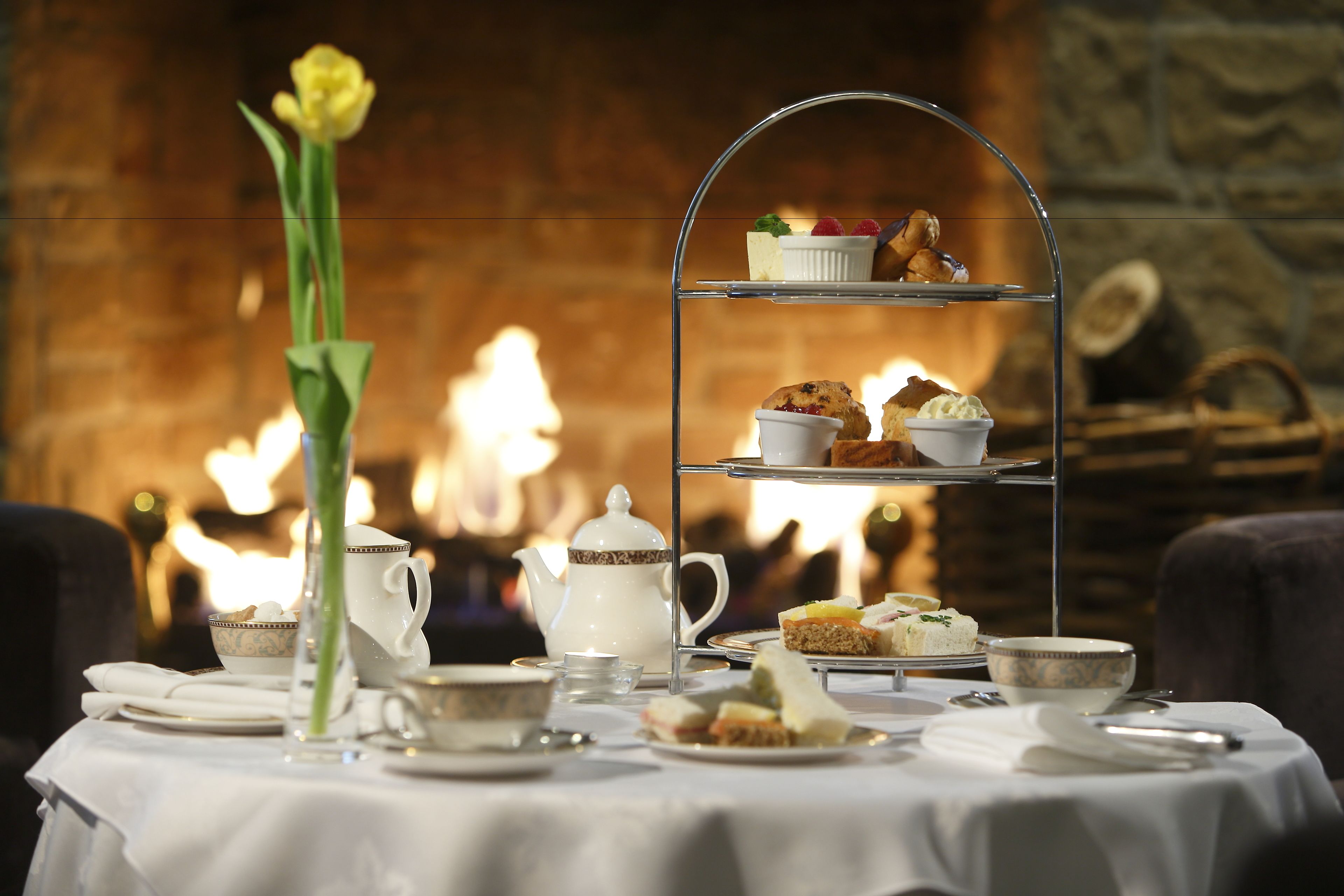 A table set for afternoon tea with a tiered tray, teapot, cups, and a vase of flowers.