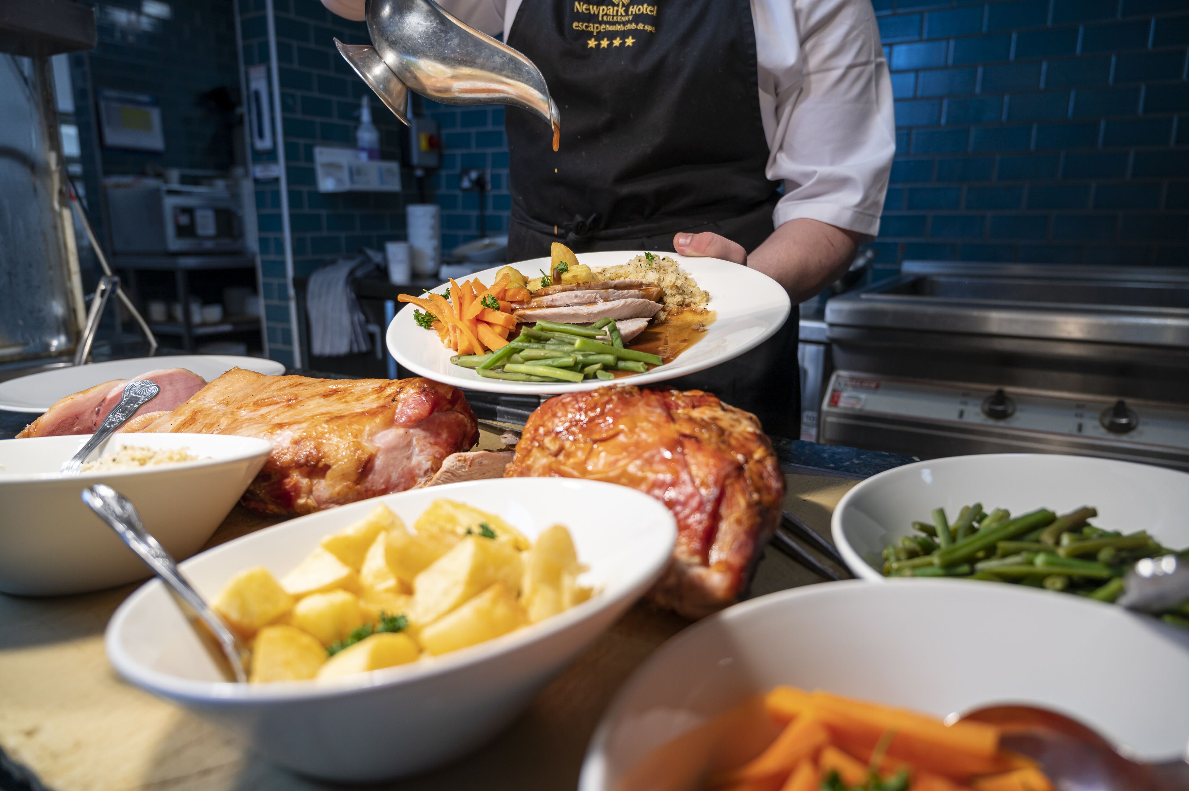 Un chef versant une sauce sur une assiette de nourriture, comprenant de la viande et des légumes, dans une cuisine.