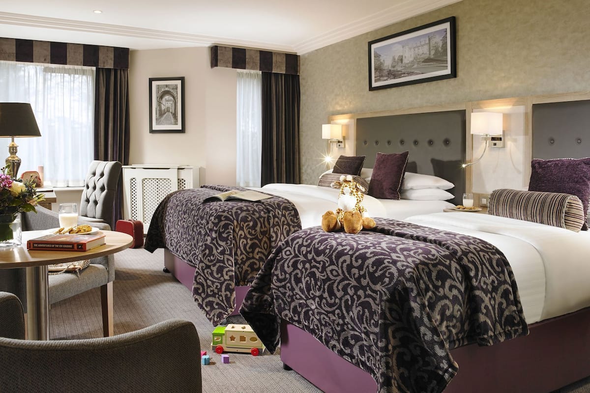 Una habitación de hotel con una cama grande, dos mesitas de noche, una silla, una pequeña mesa con lámpara y un cuadro en la pared.